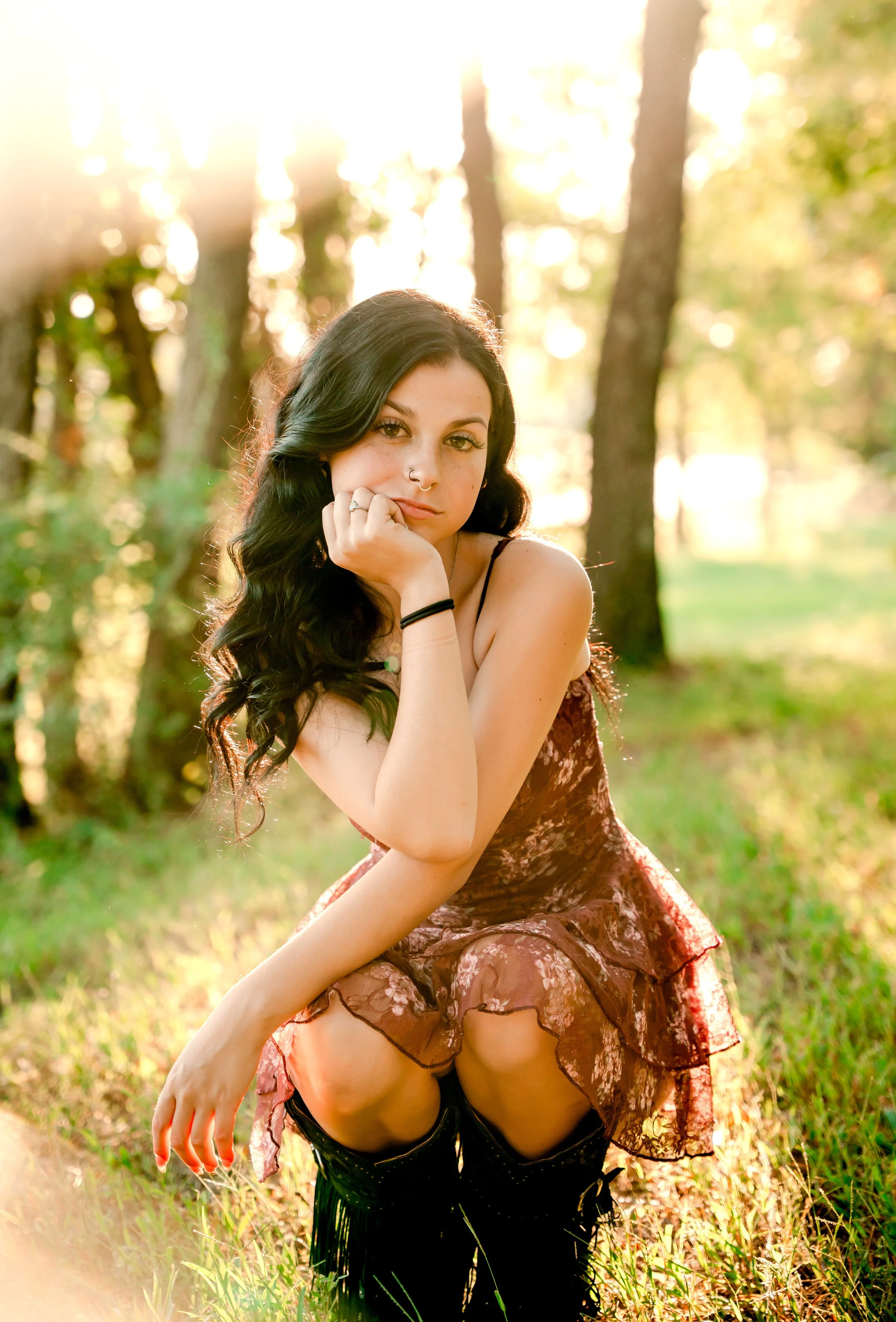 A young woman with dark wavy hair and light skin, sitting outdoors in a wooded area with sunlight filtering through the trees, resting her chin on her hand and wearing a floral dress and black boots.