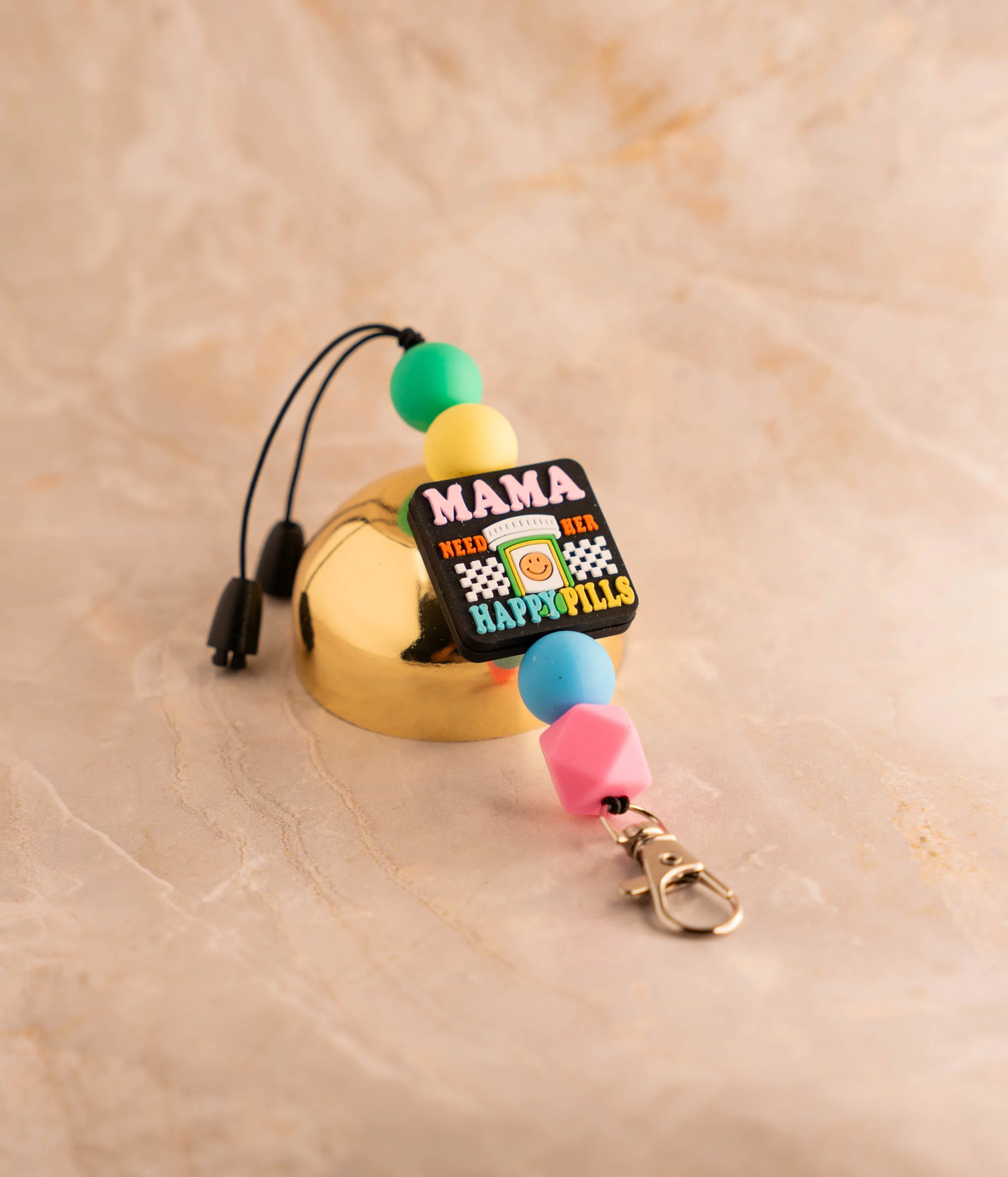 Colorful beaded keychain with a black square pendant that says "MAMA NEED HER HAPPY PILLS" and a smiling face, placed on a shiny gold heart-shaped object on a beige marble surface.