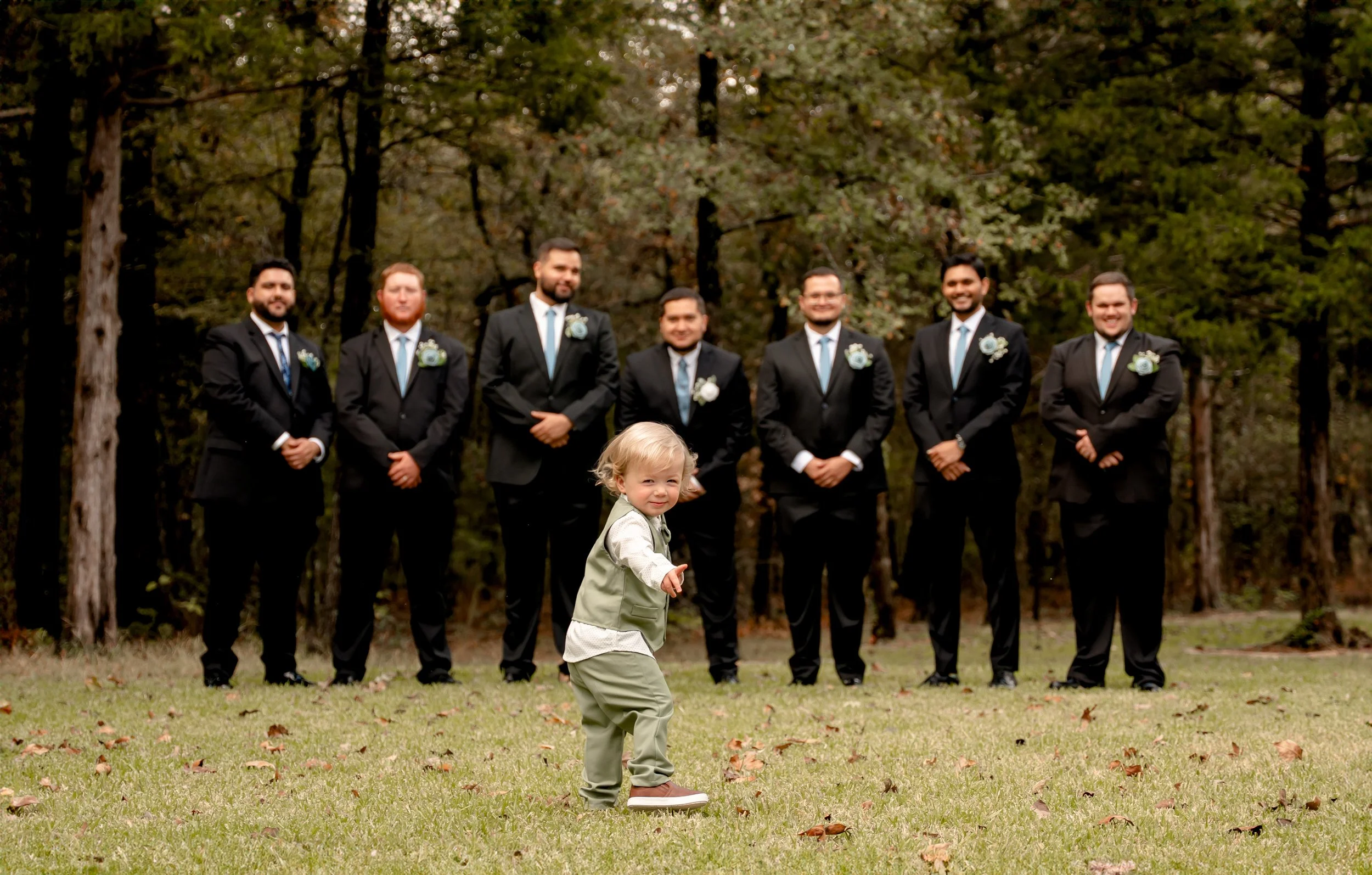 A young child standing on grass in front of seven men dressed in black suits with blue ties and boutonnieres, outdoors in a wooded area