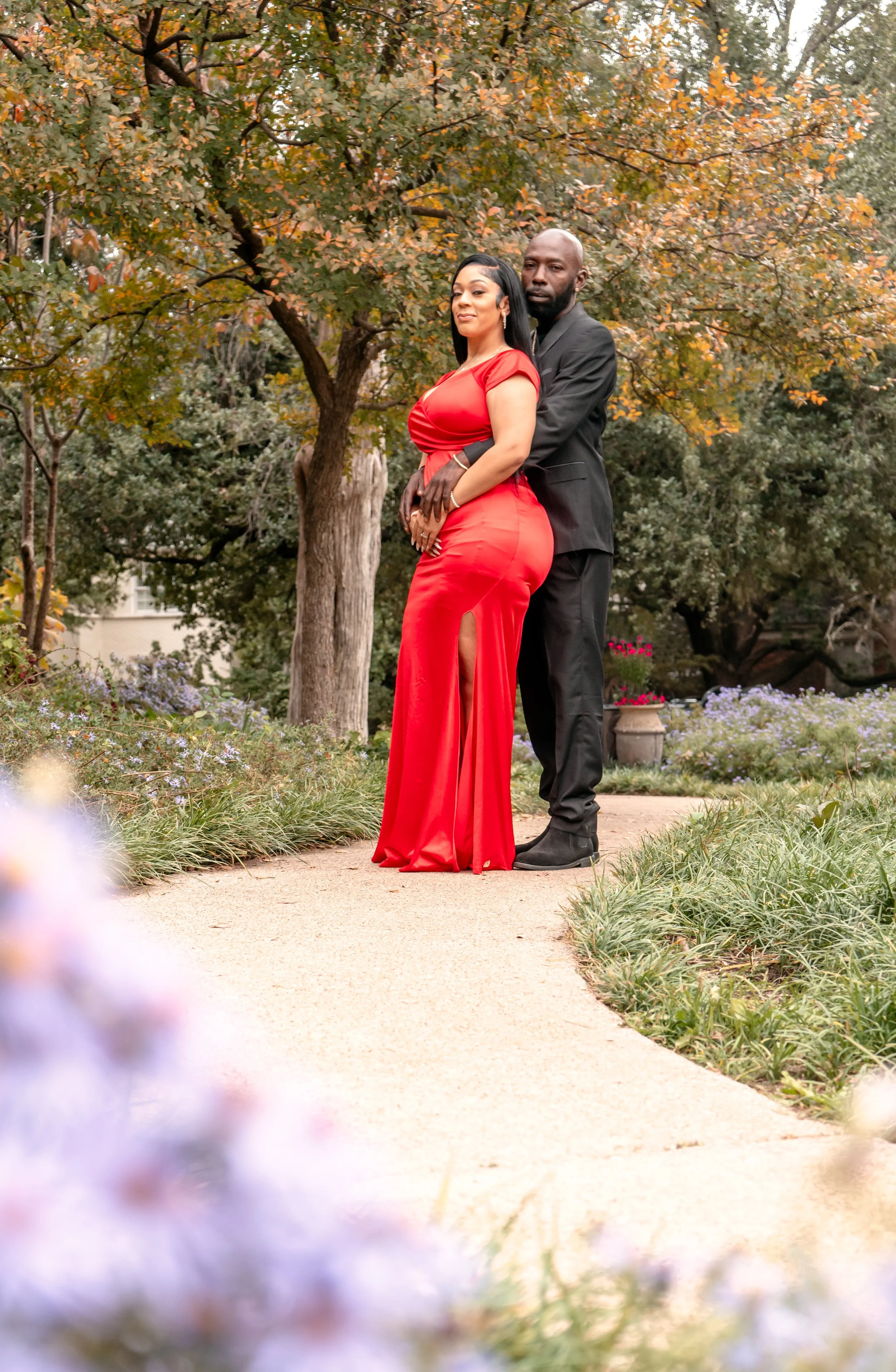 A couple dressed in formal attire standing closely together on a park pathway with trees and flowers in the background during fall.