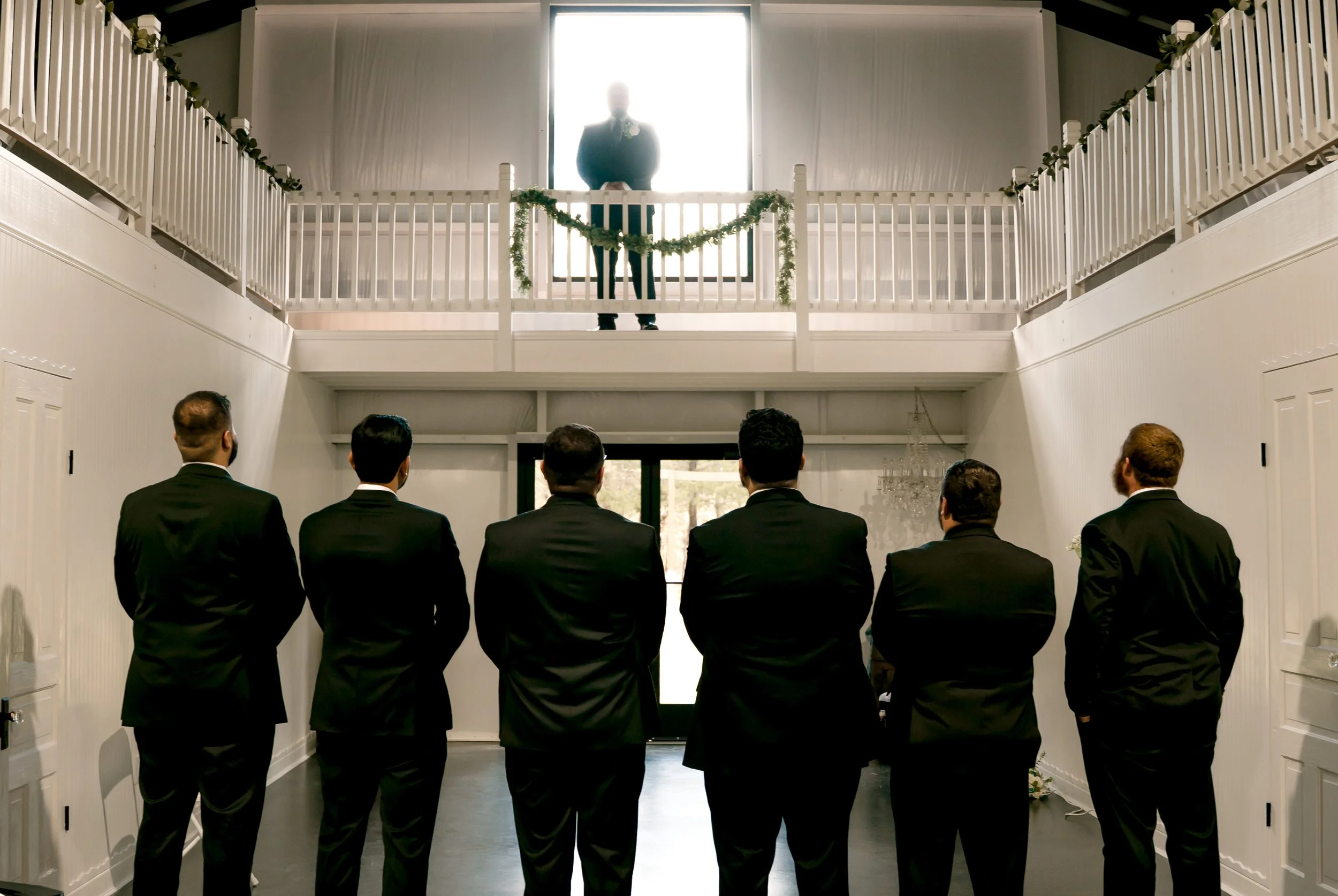Group of six men dressed in black tuxedos, standing inside a white venue, watching a man stand on a balcony above with bright light behind him. The venue has white walls, a black door, and a balcony with white railings decorated with green garland.
