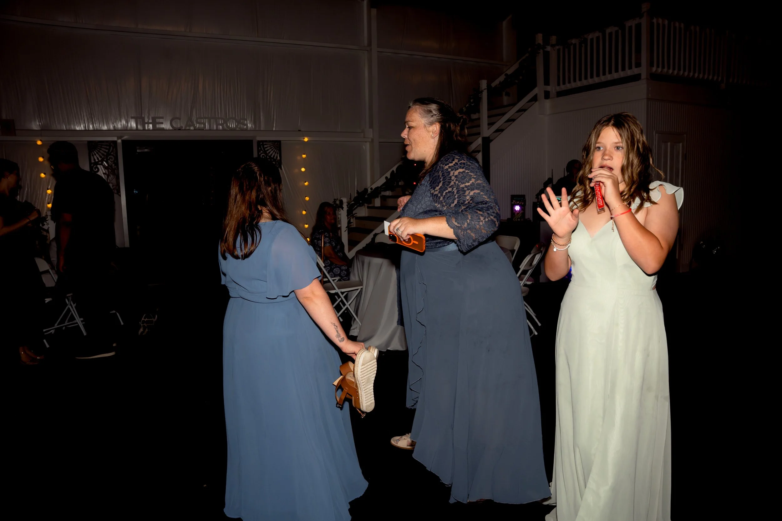 A woman wearing a navy lace top and long skirt appears to be dancing with a girl in a blue dress, while another girl in a white dress looks on and holds a snack, at an indoor gathering.