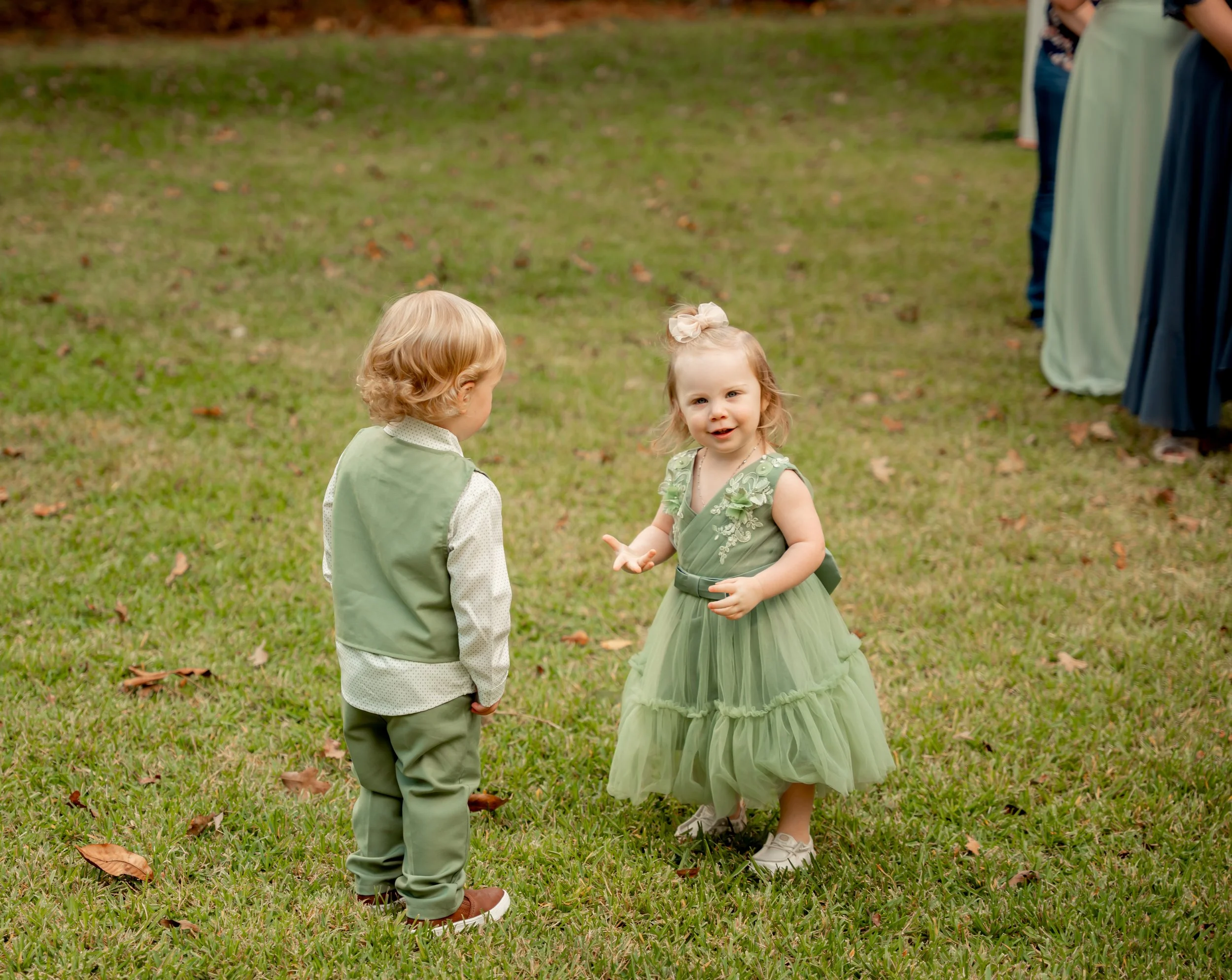 A young girl and boy dressed in green outfits standing on a grassy field, with the girl speaking to the boy.