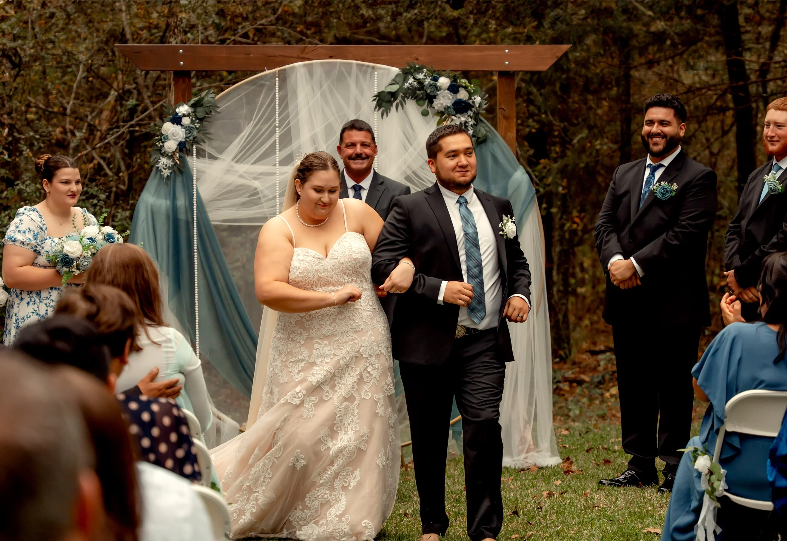 Outdoor wedding ceremony with bride and groom walking arm in arm beneath decorative arch, surrounded by wedding party and seated guests, wooded background.