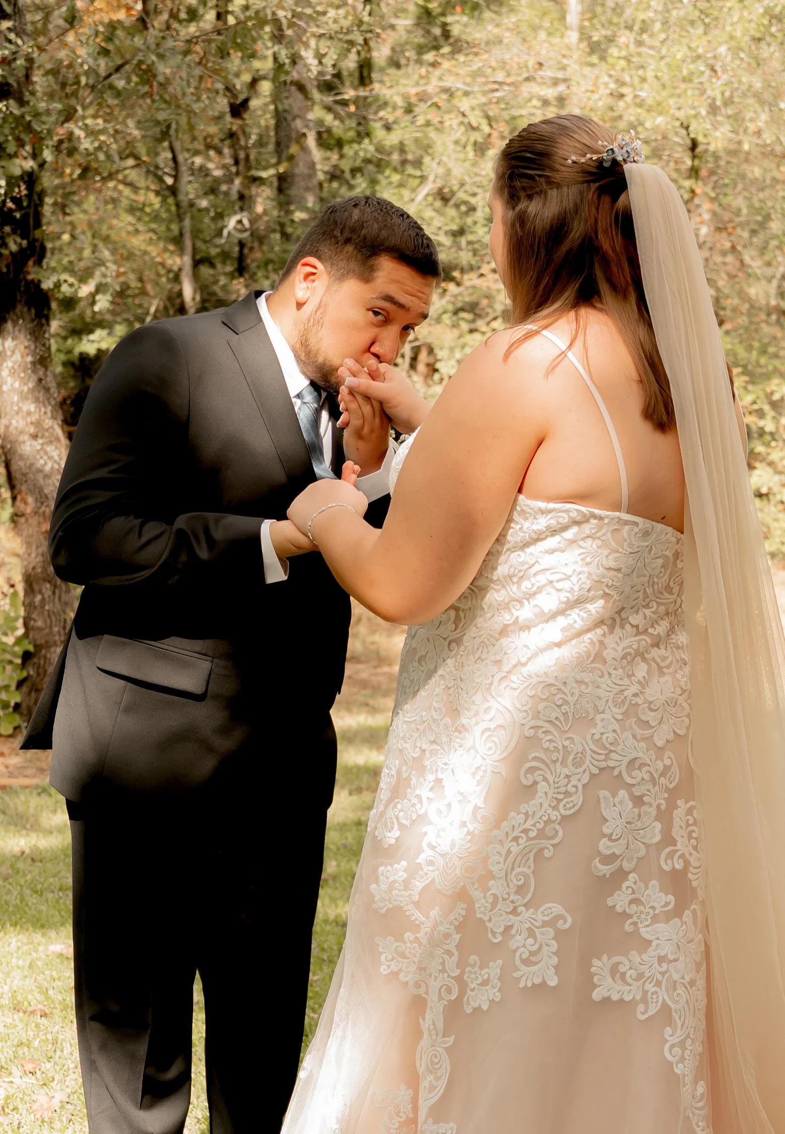 A groom in a black suit kisses the hand of a bride in a wedding dress outdoors during the day.