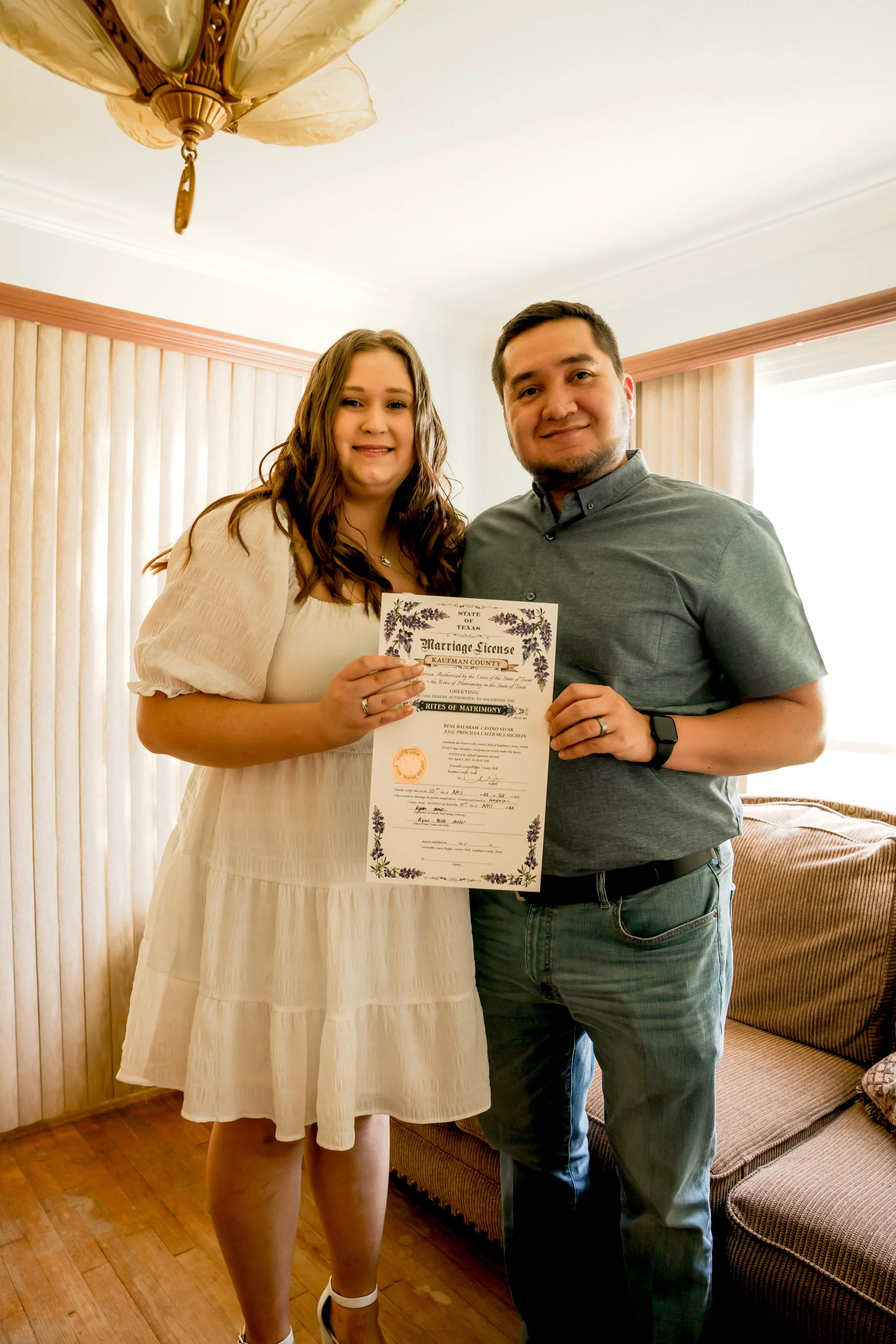 A couple holding a marriage license and smiling indoors during daytime. The woman wears a white dress, and the man wears a gray shirt. They are standing near a window with vertical blinds and a couch in the background.