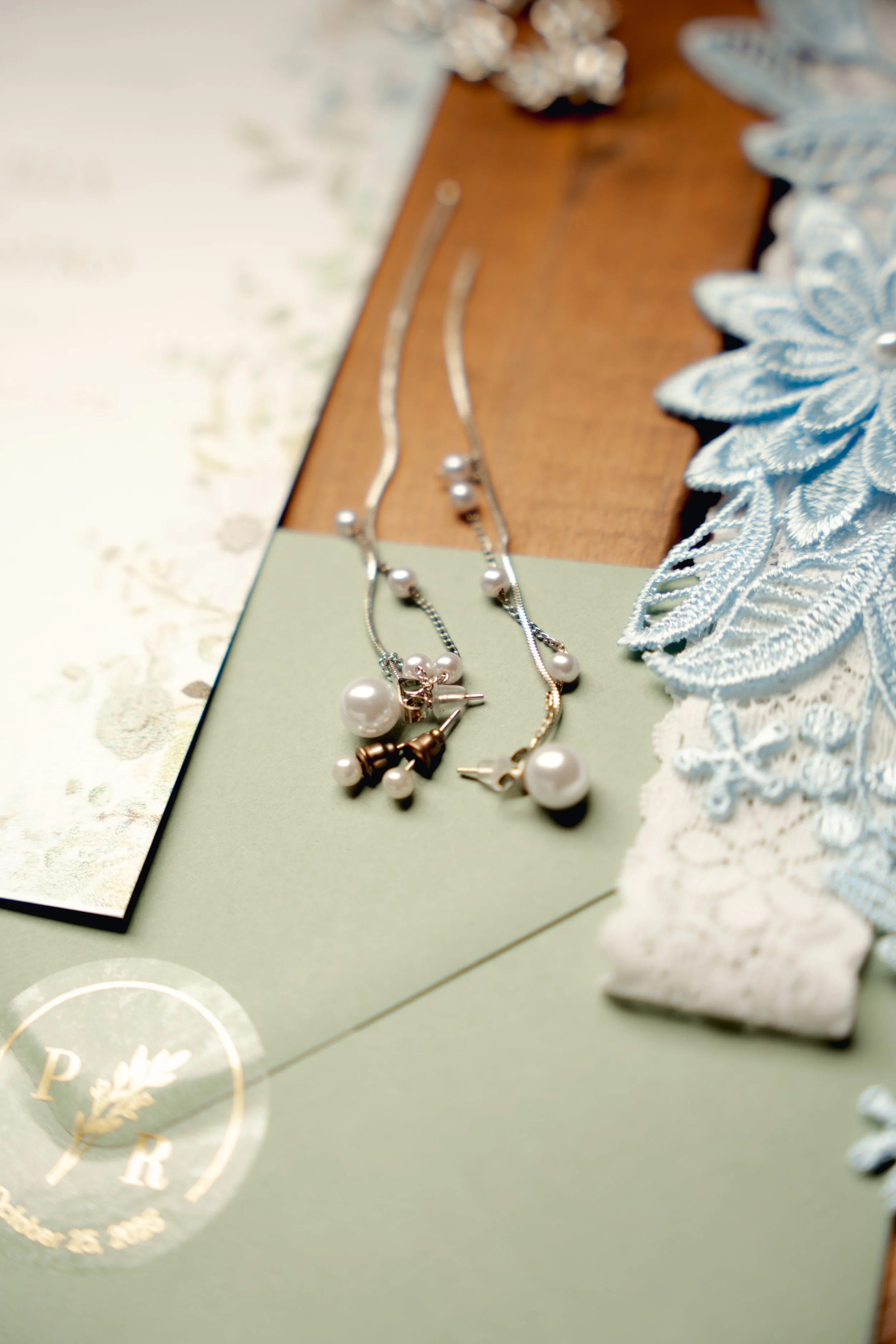 Silver and gold jewelry, including pearl necklaces and earrings, arranged on a green and wooden surface with lace fabric on the right.