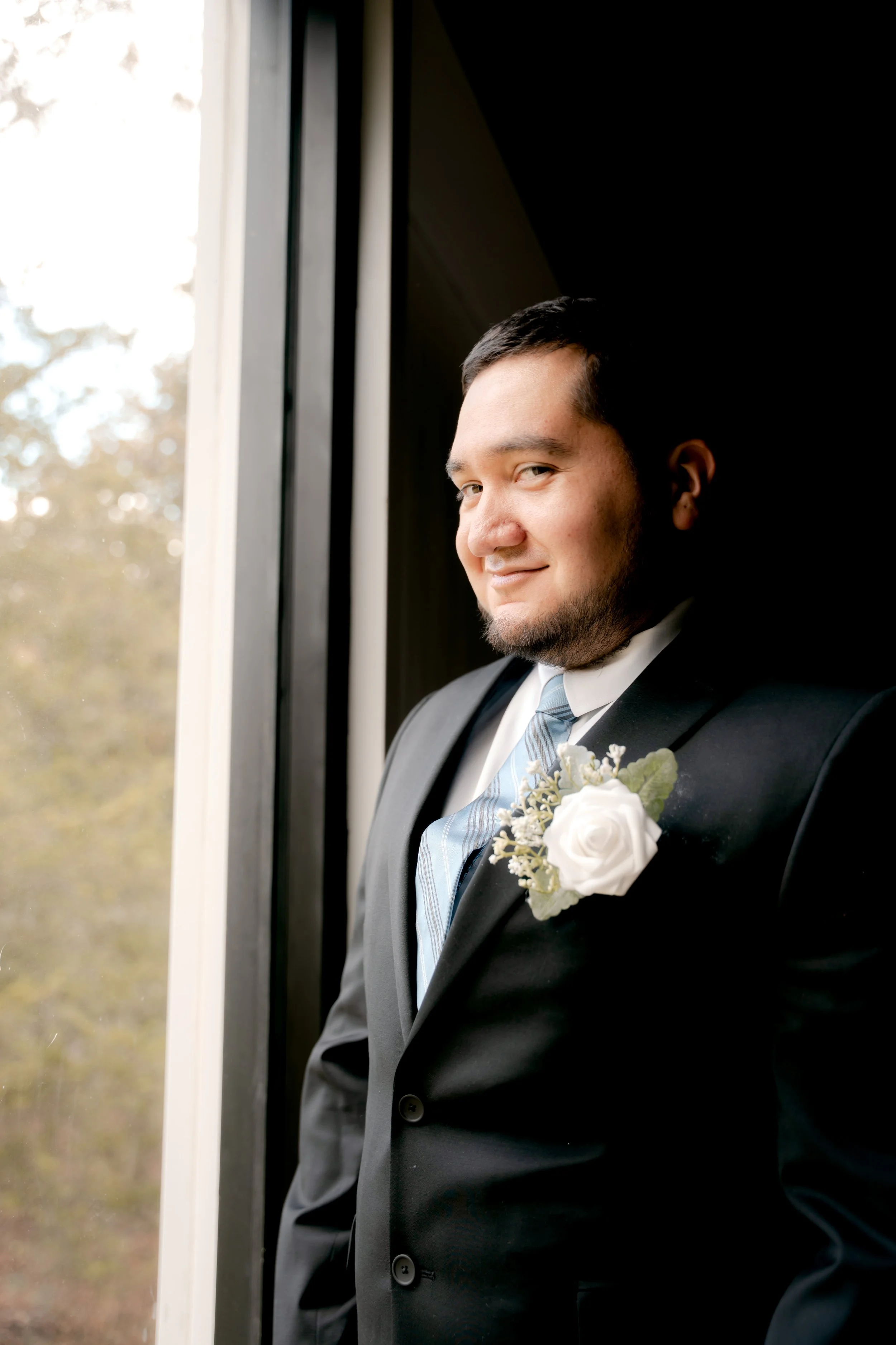 A man in a black suit with a white shirt and a striped tie, standing near a window with a white flower boutonniere on his lapel, smiling at the camera.