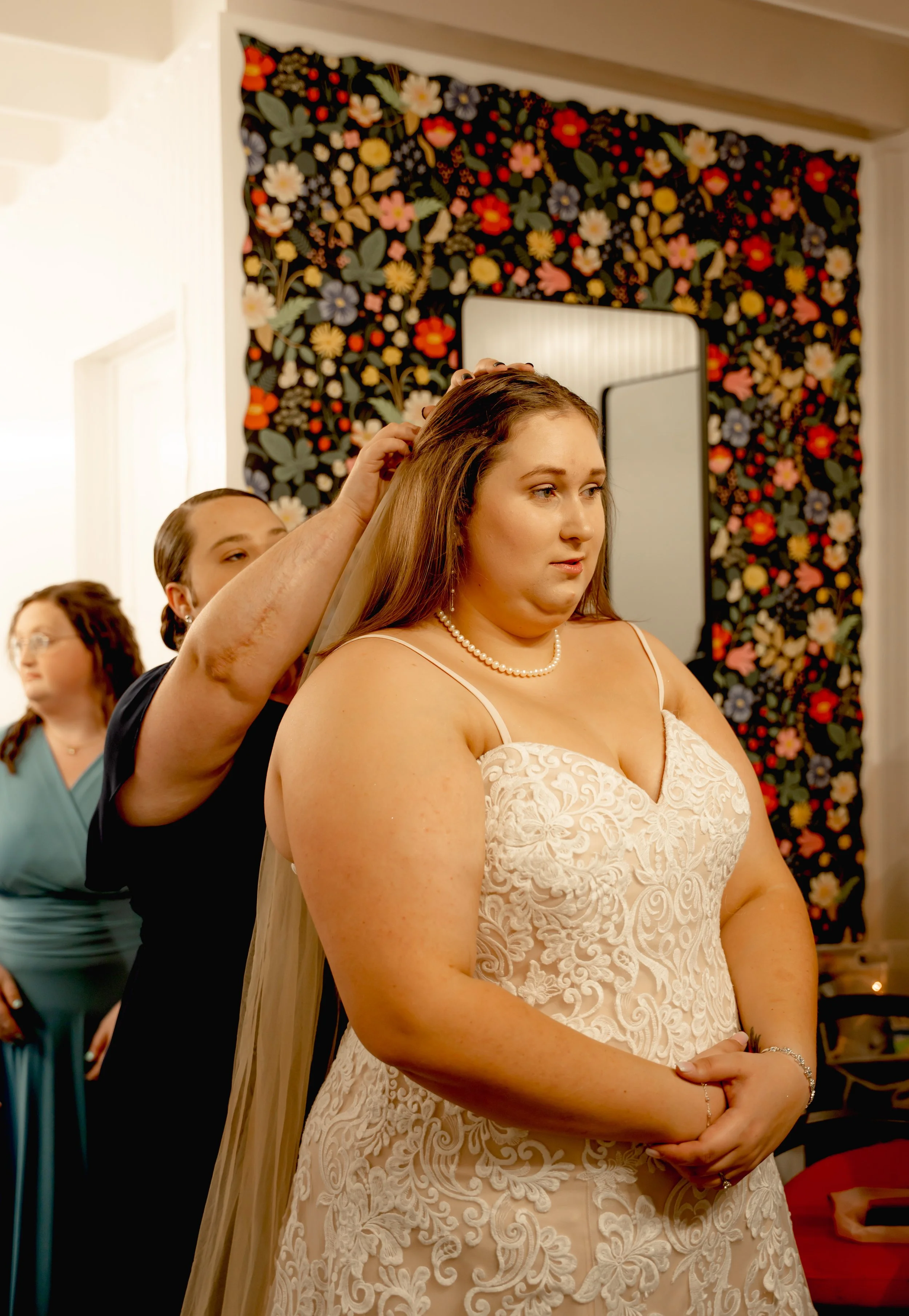 A bride getting ready with help from her attendant, standing in front of a floral patterned curtain, wearing a white dress and pearl necklace.
