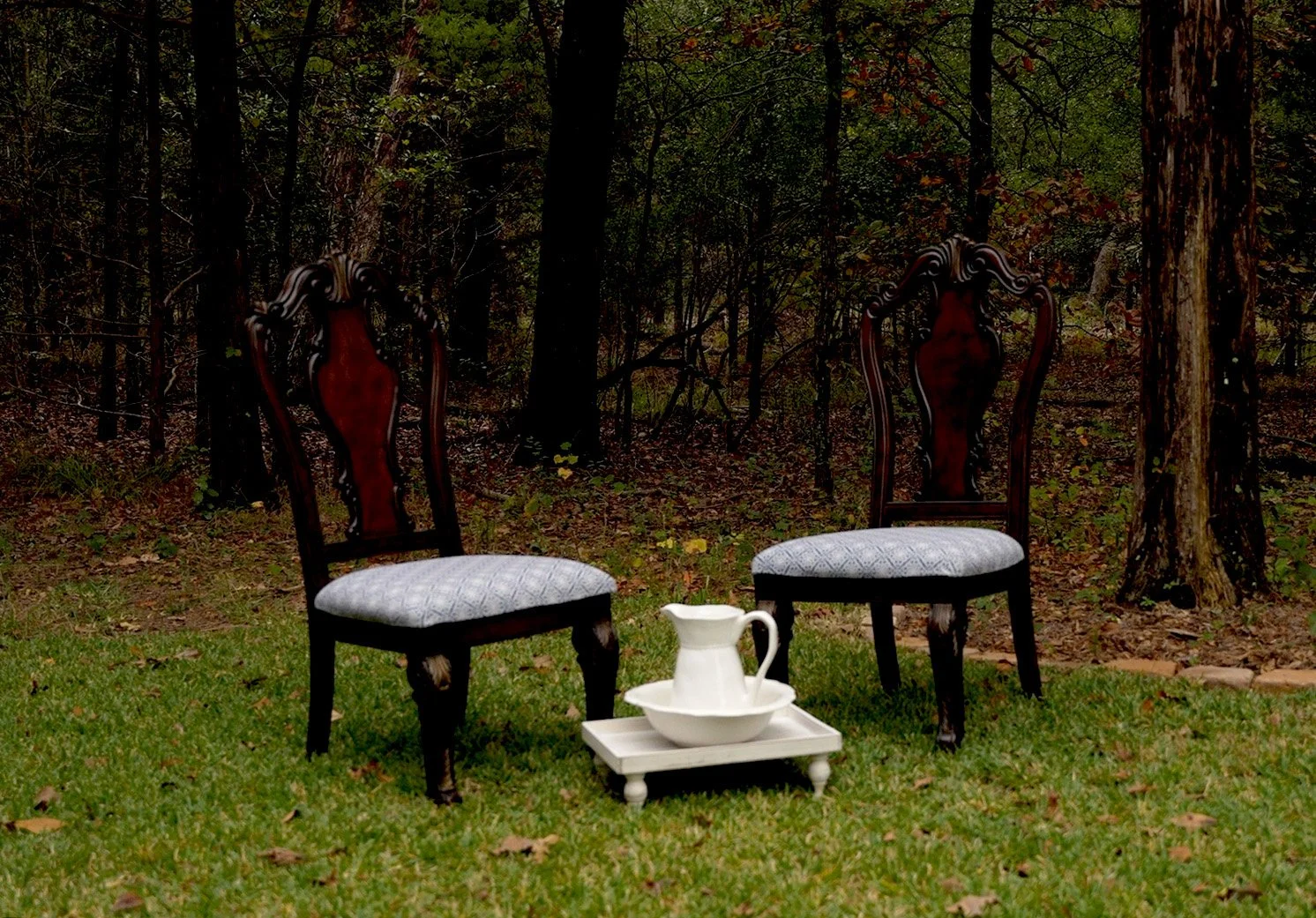 Two vintage wooden chairs with upholstered seats are positioned outdoors on a grassy area, with a white ceramic pitcher and bowl set on a small white tray in between them. A wooded forest with trees and foliage is in the background.