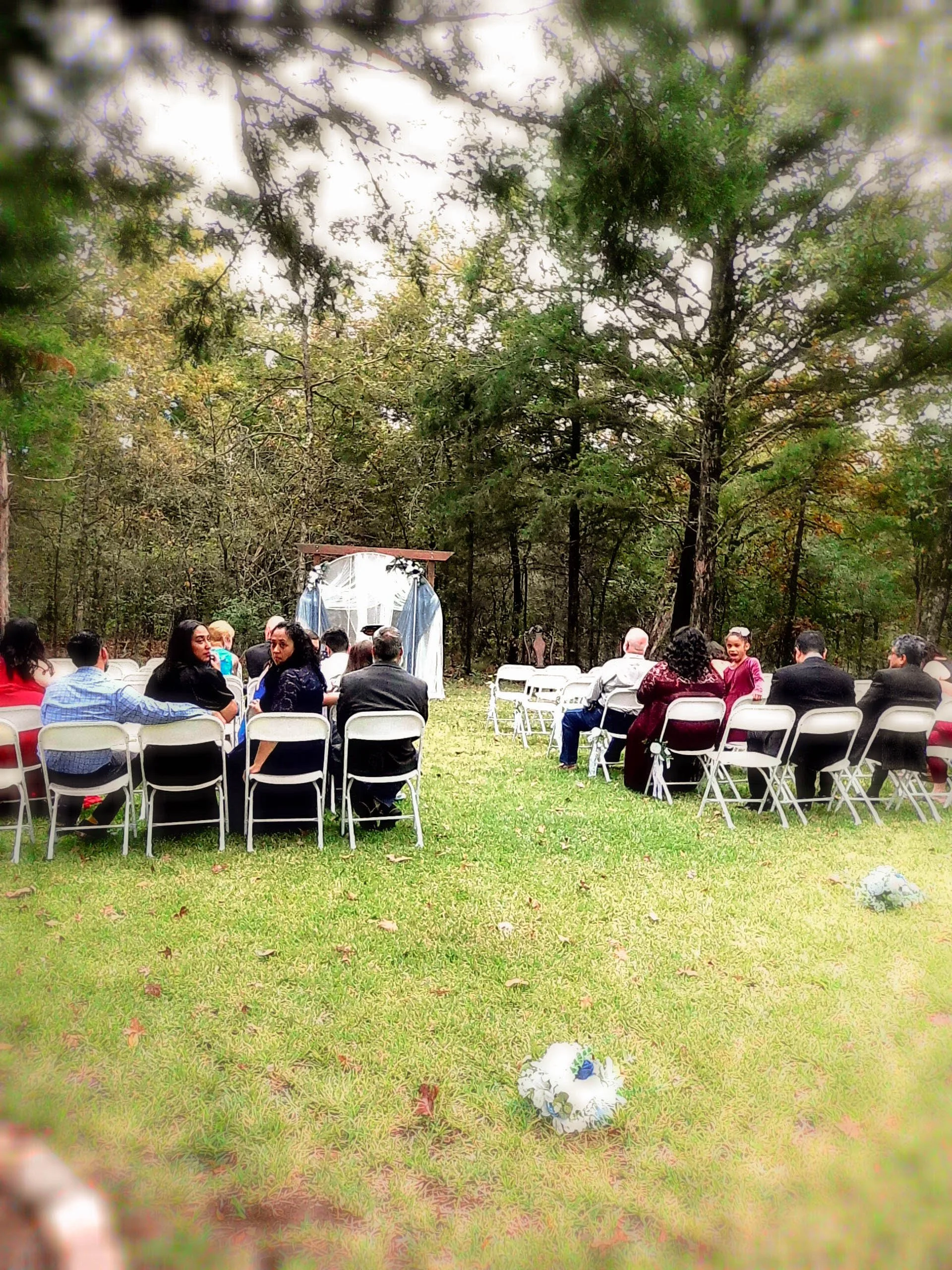 Outdoor wedding ceremony in a forested area with guests seated on white chairs, some wearing formal attire. There is a small altar with drapes in the background. The ground is grassy, and there are floral decorations on the grass and chairs.