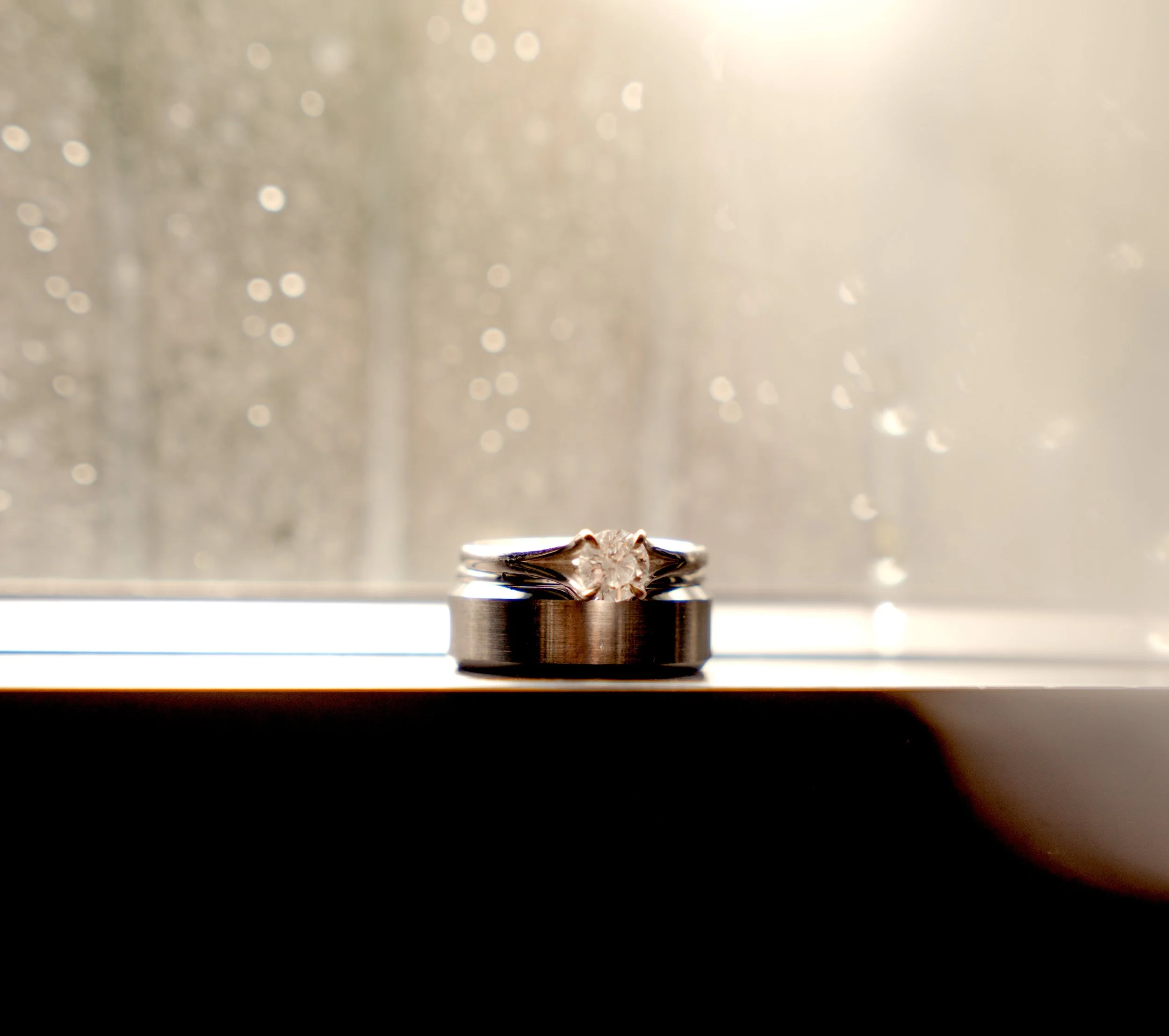 A silver ring with a large central gemstone, likely a diamond, resting on a flat surface in front of a window with rain droplets.