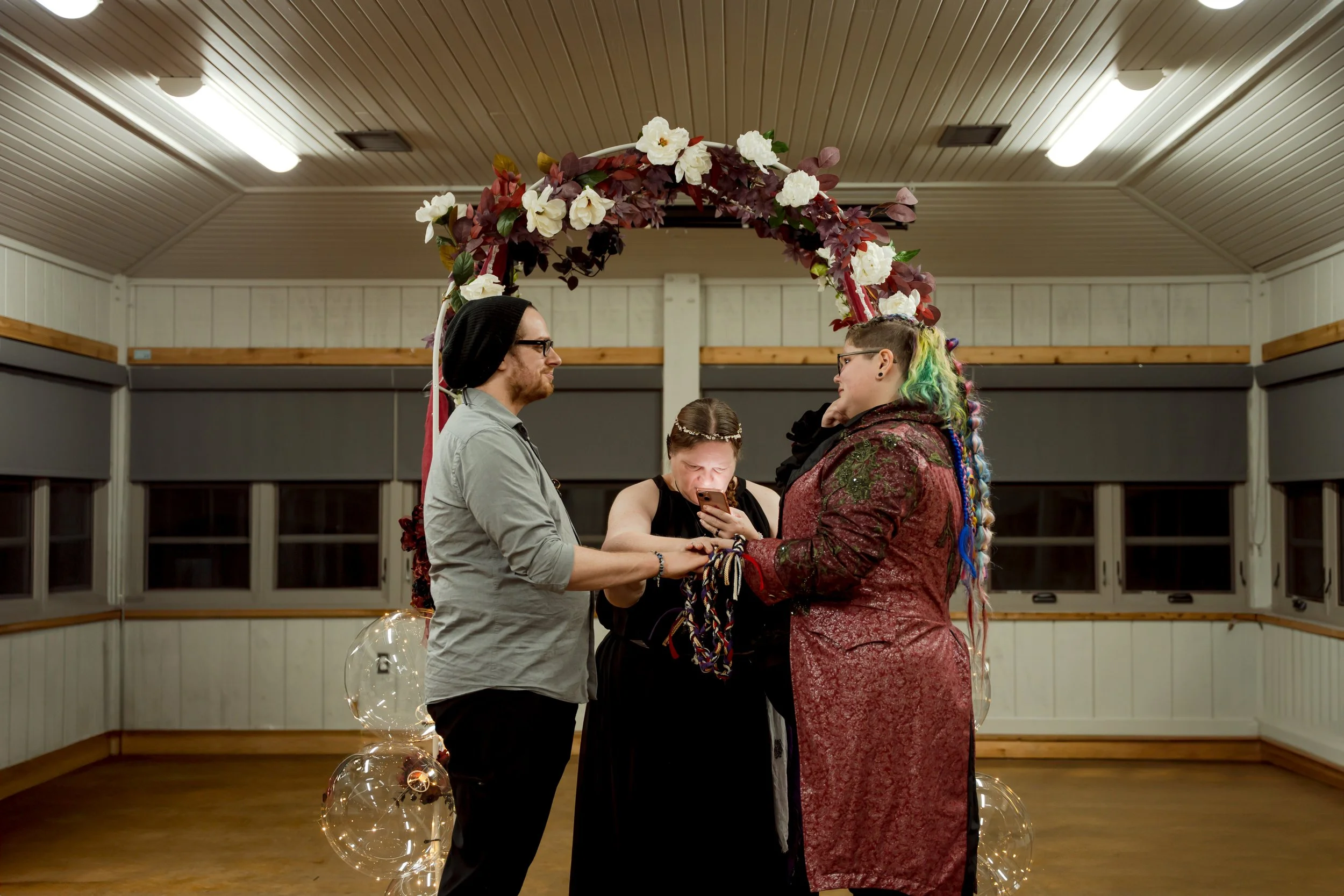 A same-sex couple and officiant during a wedding ceremony under a floral arch in a wooden-paneled room, with wedding rings, bubbles, and one person looking at a phone.