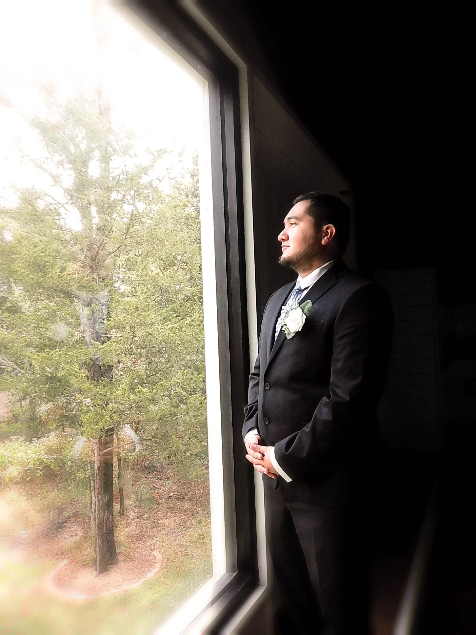 A man in a black suit and tie, with a white boutonniere, standing inside near a large window, looking out at a tree.