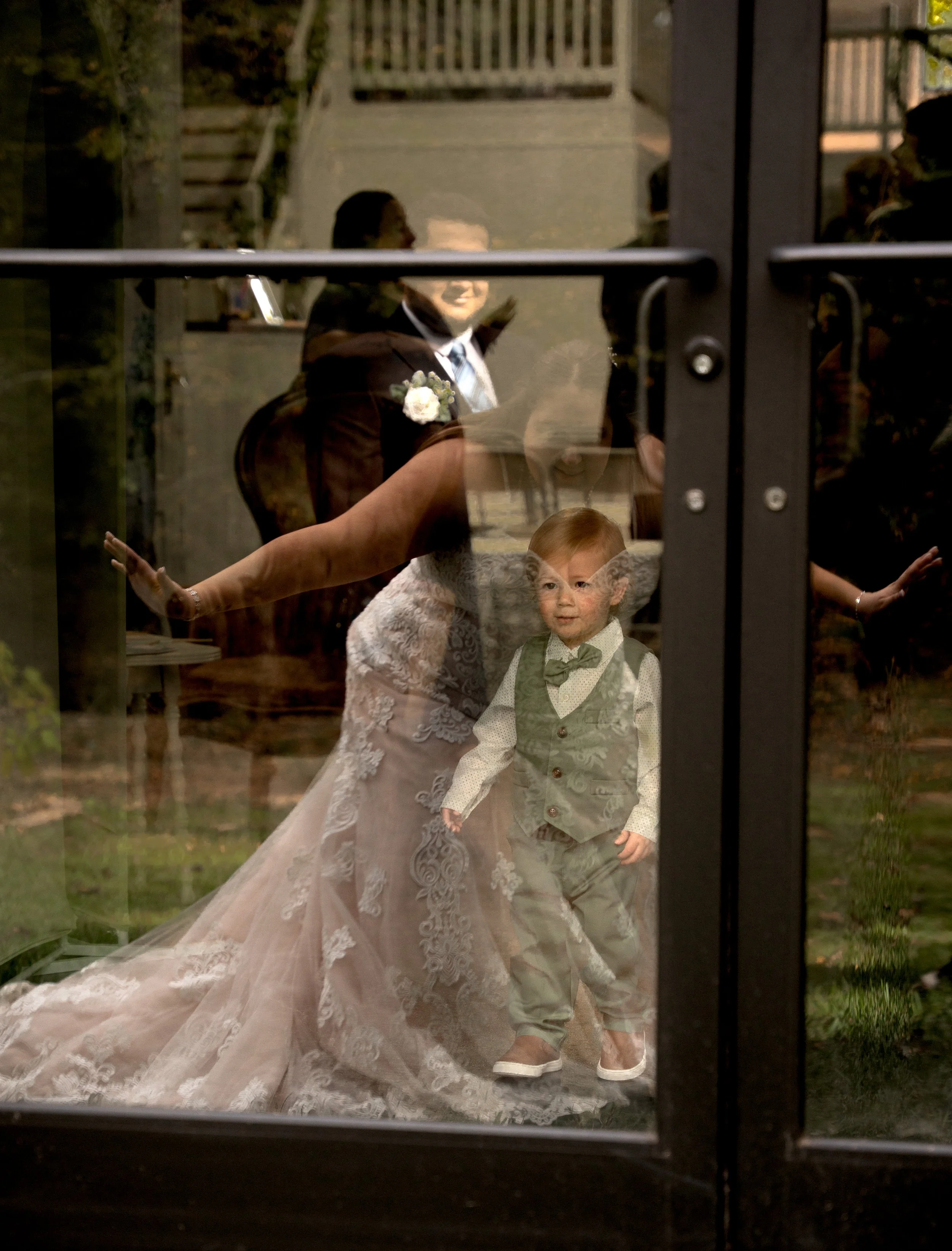Reflected image of a bride and groom, with a child in formal attire, seen through glass doors at a wedding reception.
