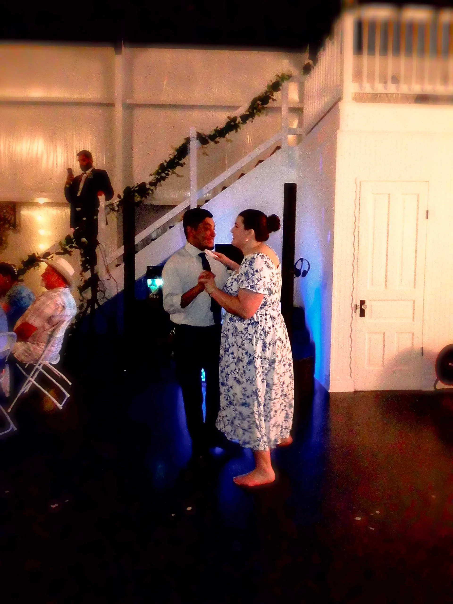 Couple dancing at a wedding reception, with a man in a white shirt and tie and a woman in a floral dress, holding hands and smiling.