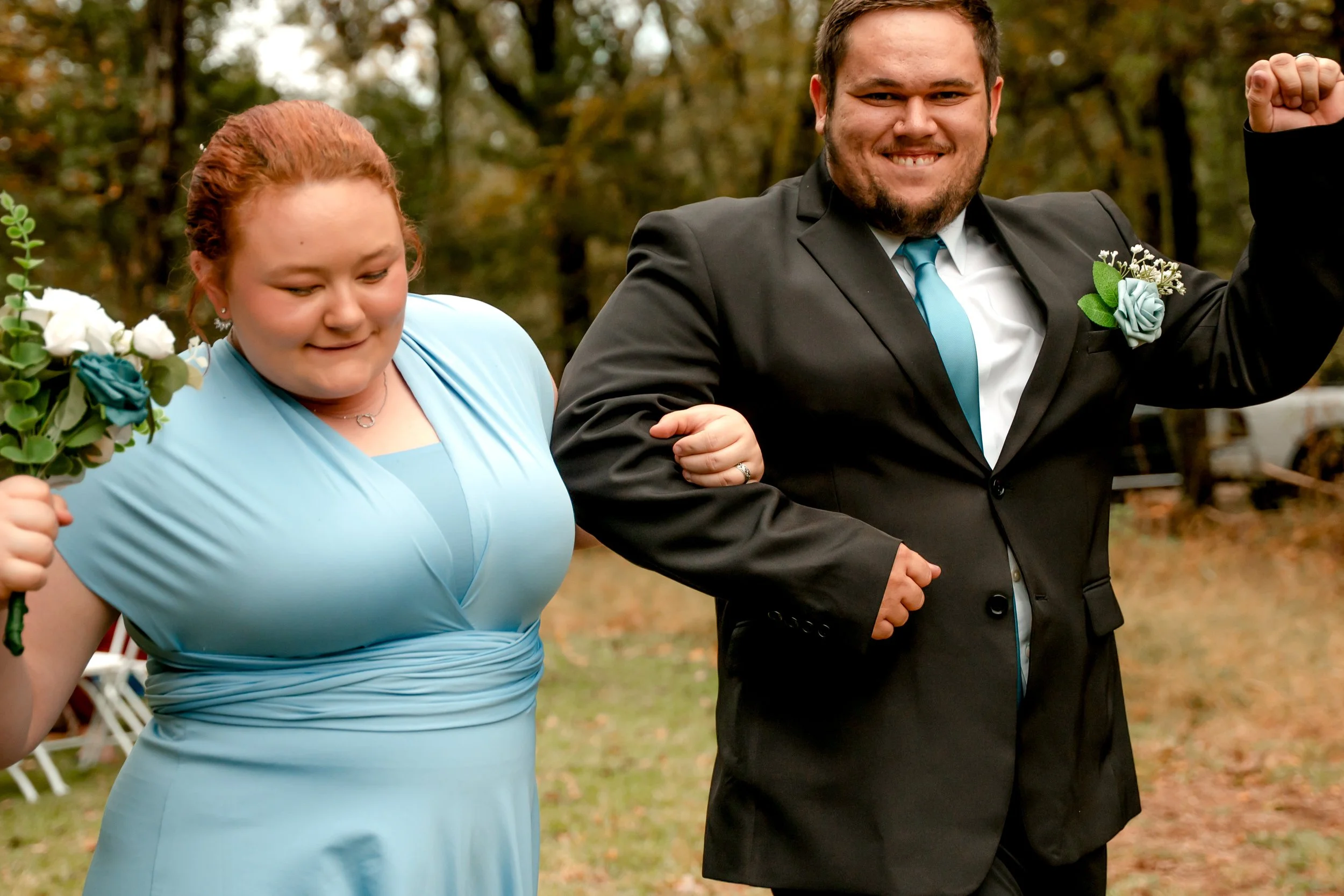 A happy couple walking outdoors at a wedding, with the woman wearing a light blue dress and the man in a black suit with a blue tie, both smiling and demonstrating confidence.