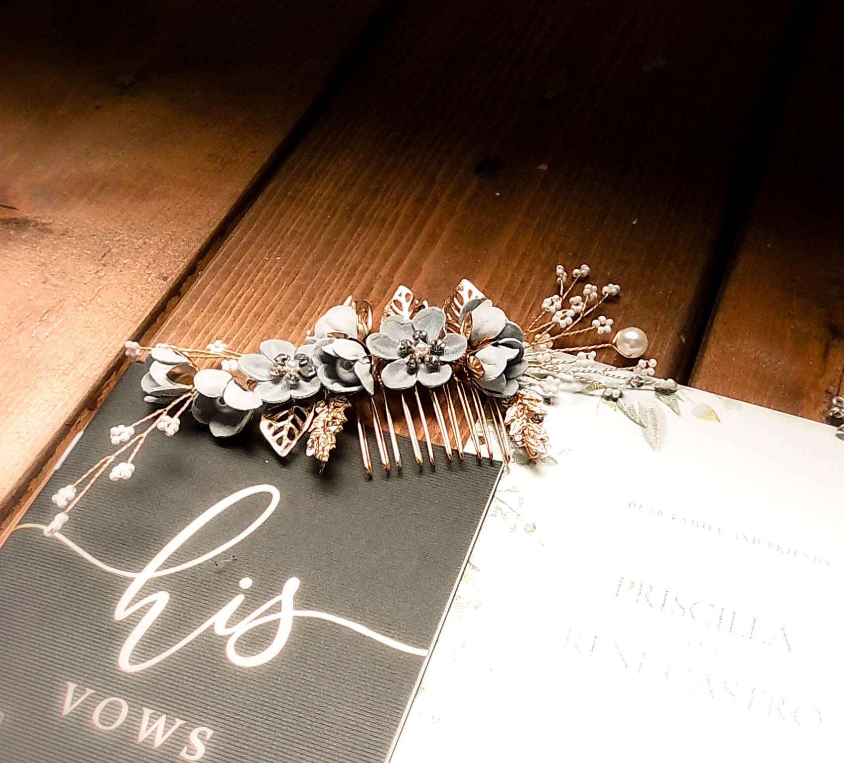 A decorative hair comb with gray flowers, gold leaves, pearls, and small white seed beads, placed on top of a black card with white cursive writing and another partially visible document, both on a wooden surface.