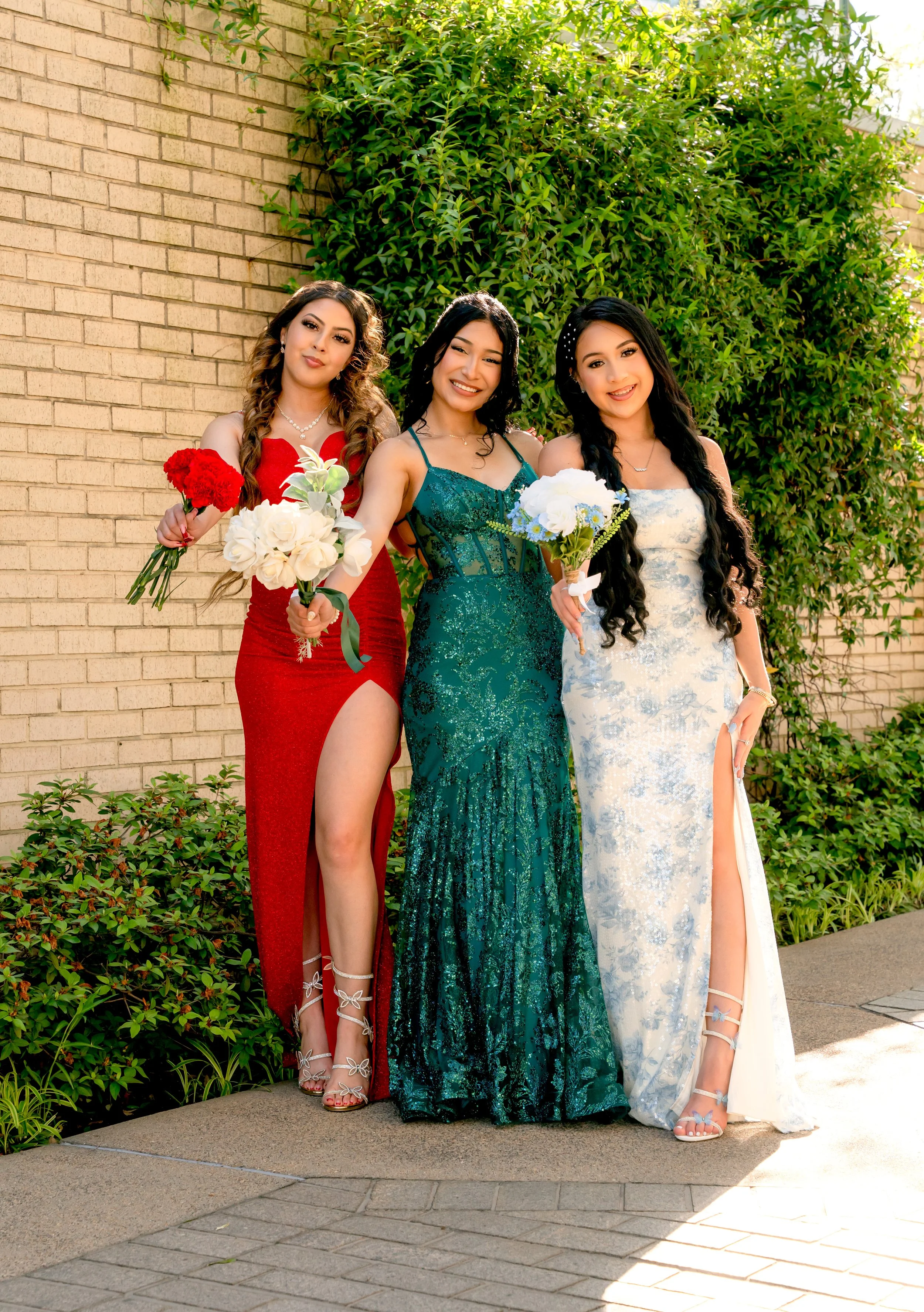 Three women in elegant dresses holding bouquets of flowers, standing outdoors against a brick wall and greenery, posing for a photo.