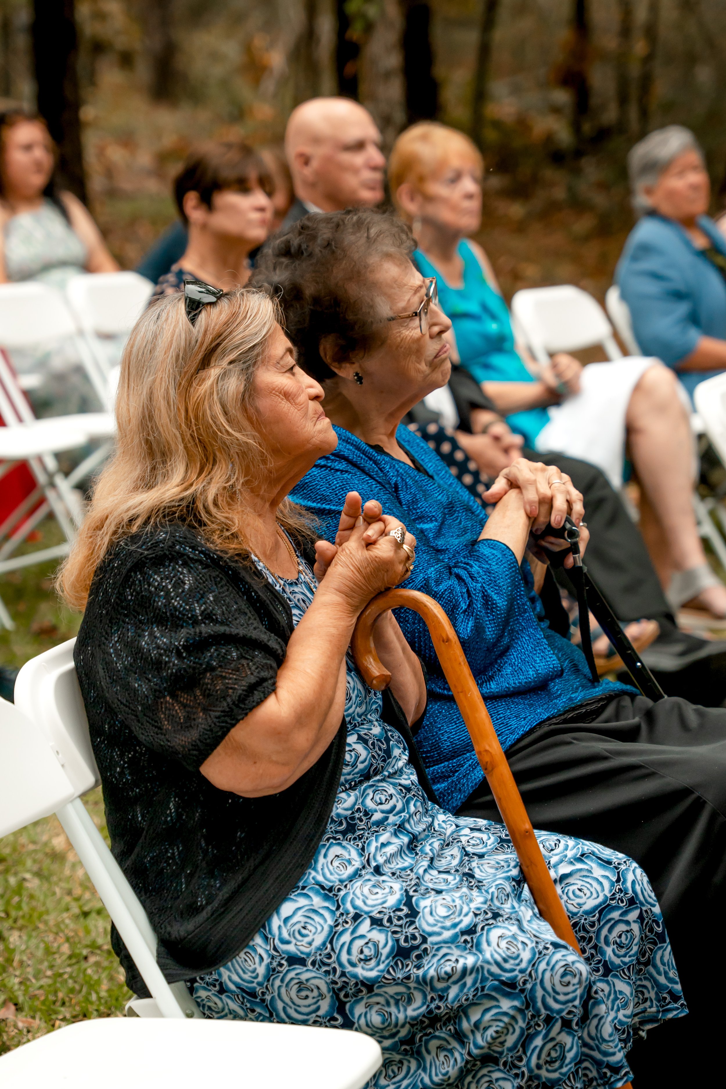 Group of elderly women and men sitting outdoors on white folding chairs, attending an event in a wooded setting.