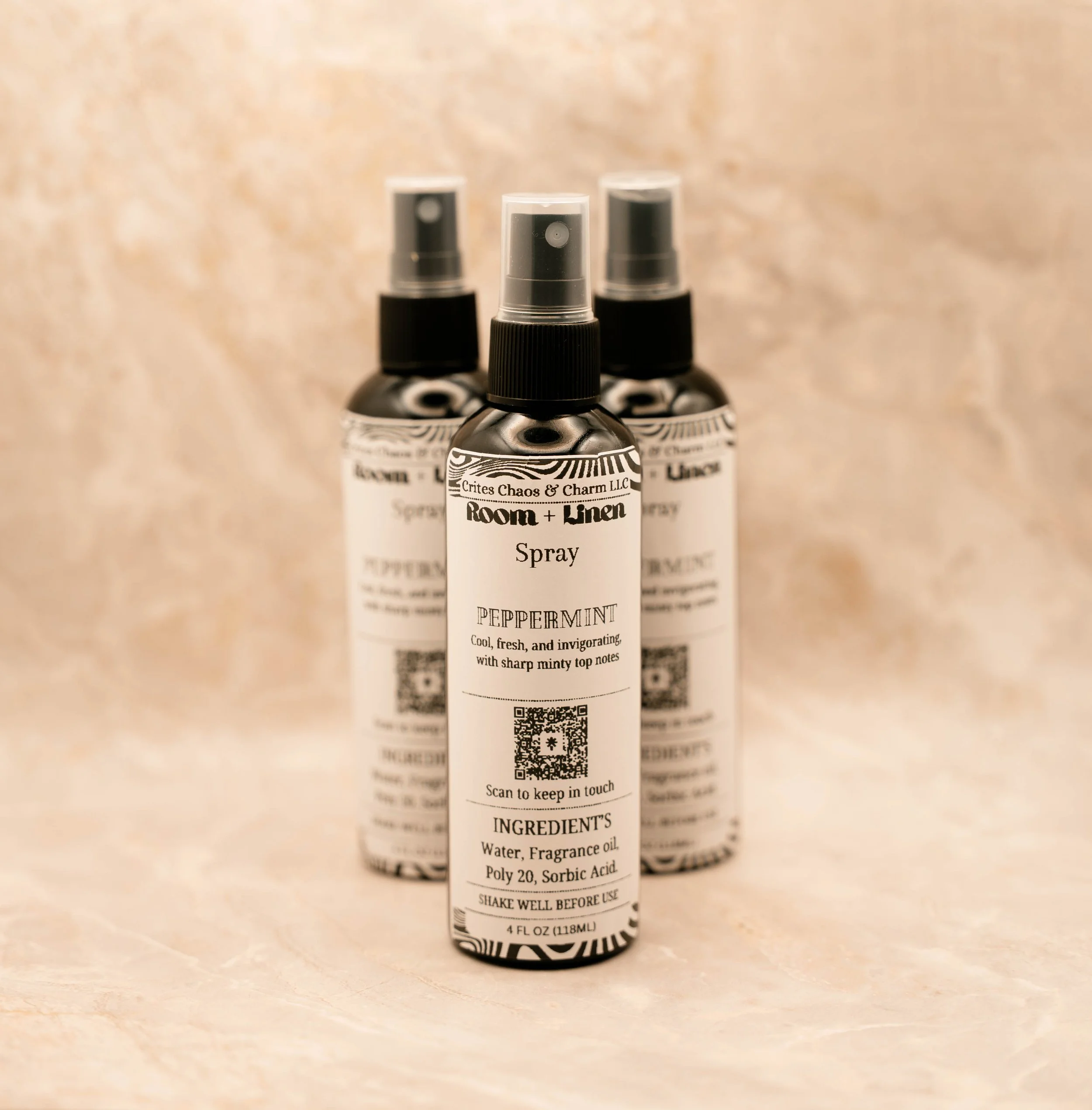 Three bottles of Room + Linen spray with peppermint scent, featuring white labels, black spray caps, and QR codes, set against a light, textured background.