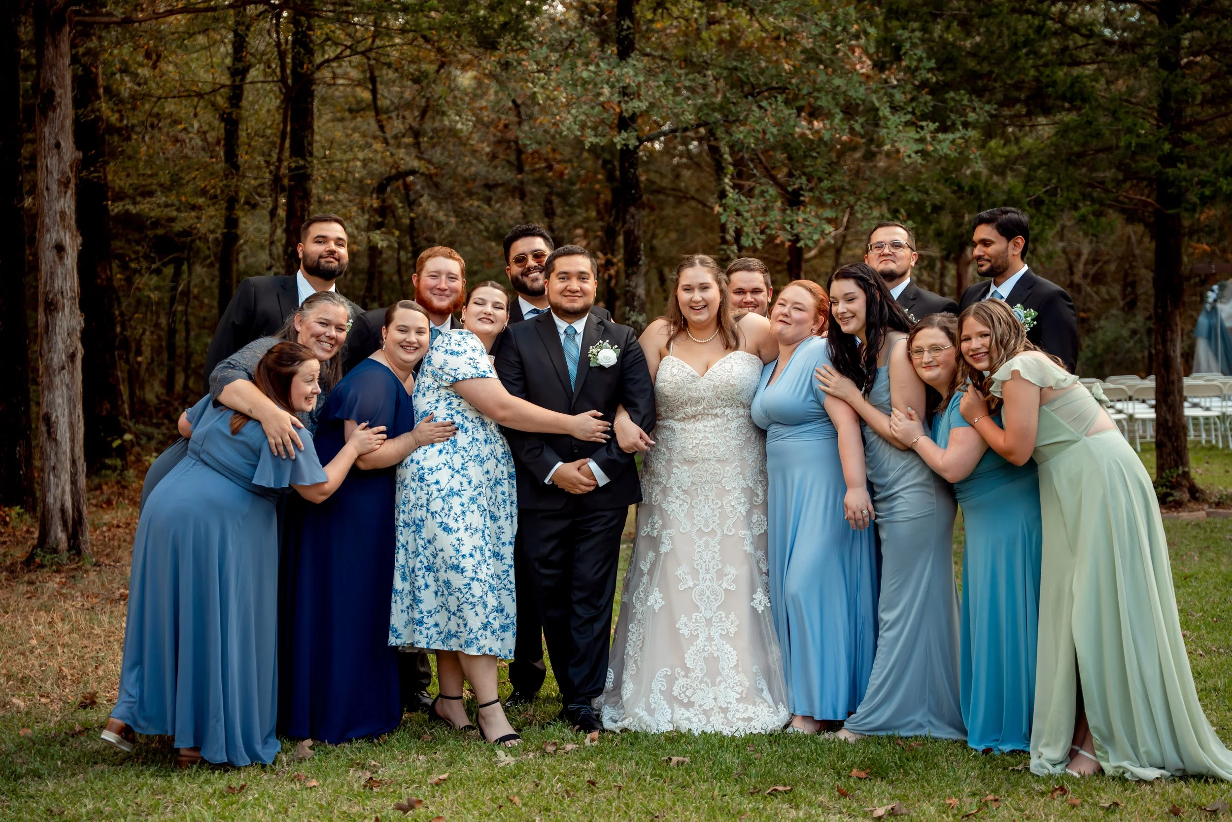 A group of people dressed in formal attire at a wedding, standing outdoors among trees, with some wearing blue dresses and others in suits, smiling and posing together.