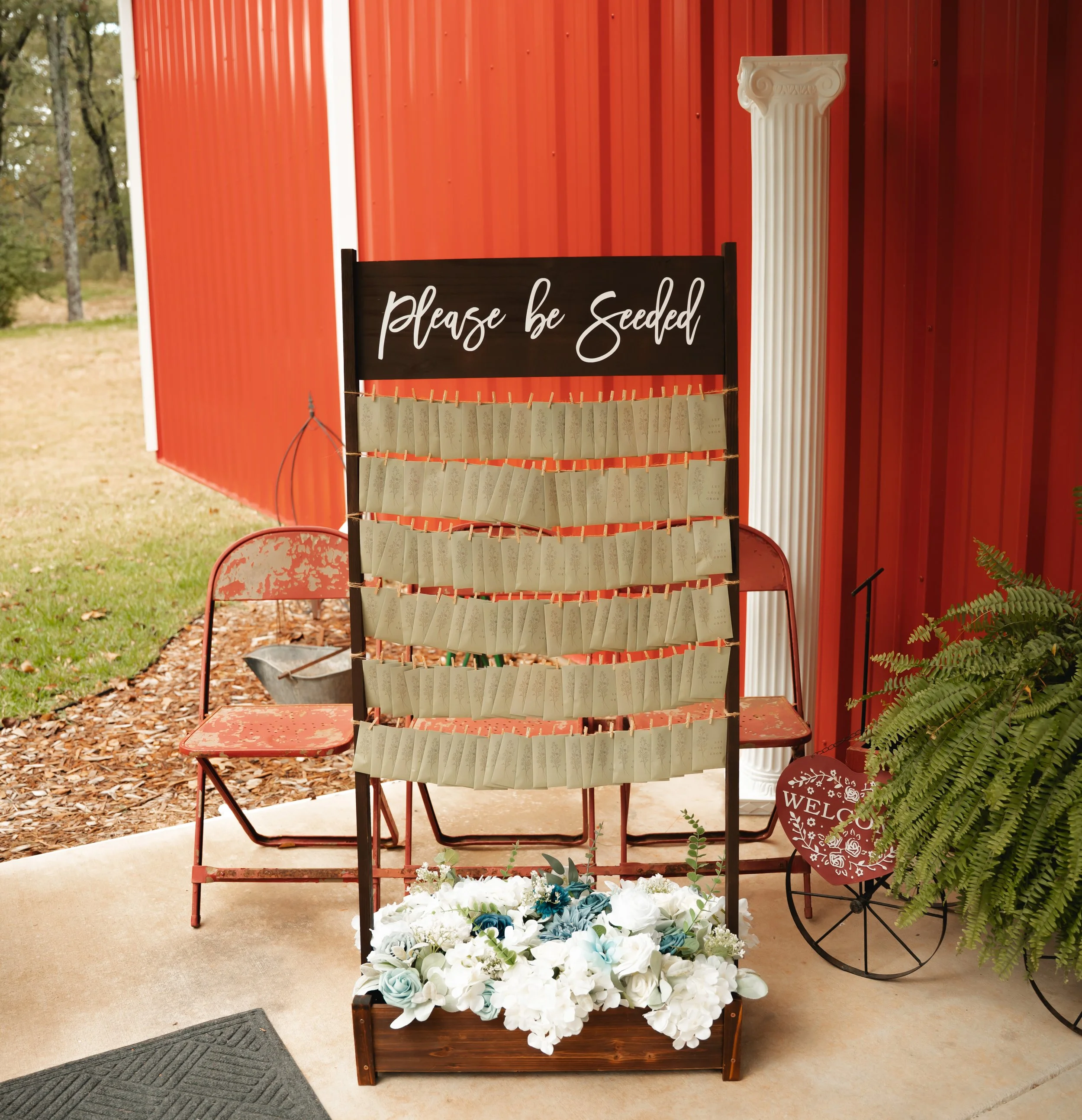 An outdoor wedding guest book display with a blackboard sign reading 'Please be Seeded,' where guests have attached paper seed packets with clothespins. The display is decorated with flowers at the base and is set against a red metal building with a 