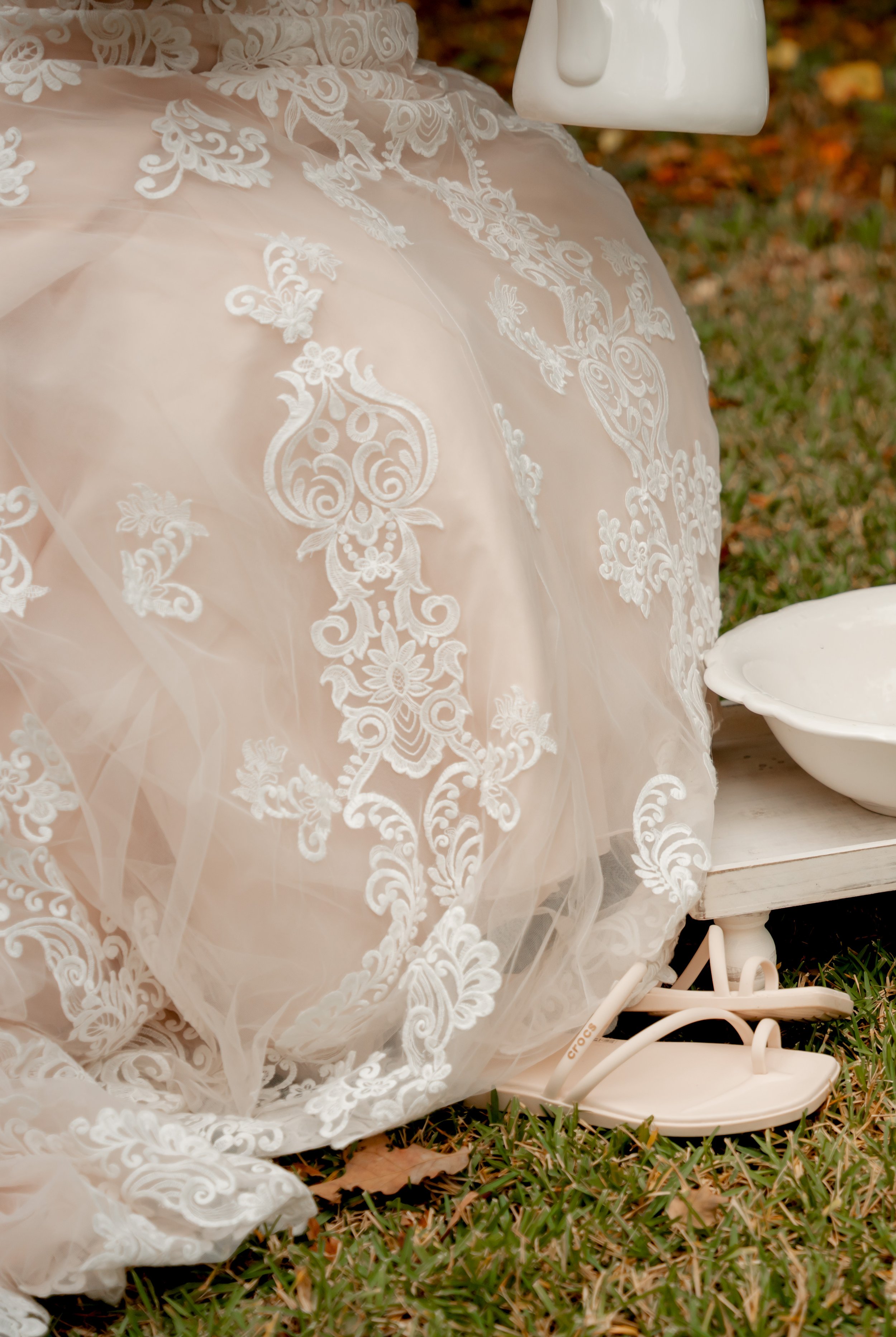 Close-up of a white lace wedding dress with intricate embroidery, lying on grass next to a small white bench and a bowl, likely part of a wedding setup.
