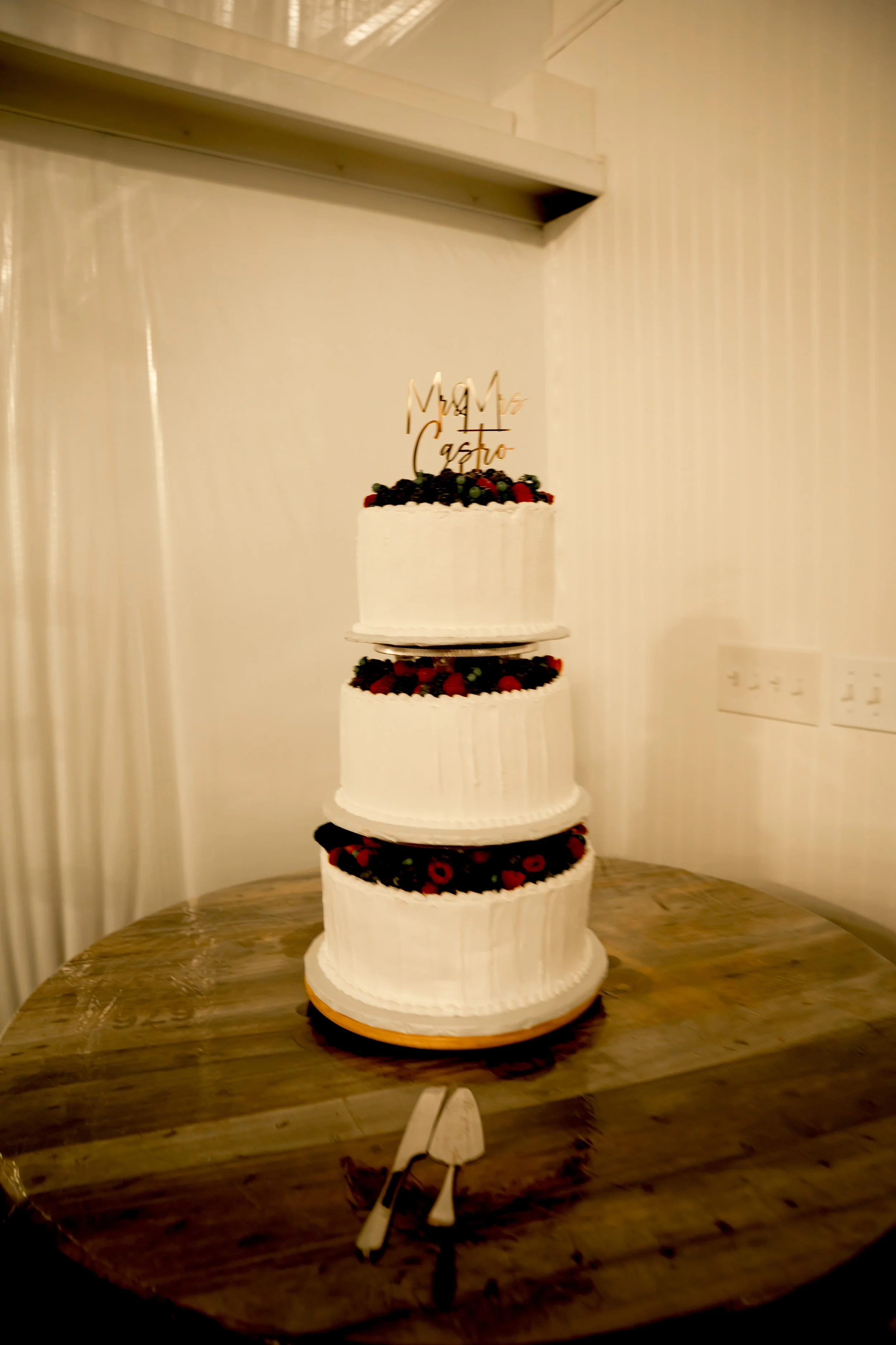A three-tiered white wedding cake decorated with berries on each layer, topped with a gold 'Mr & Mrs' cake topper, placed on a wooden table.