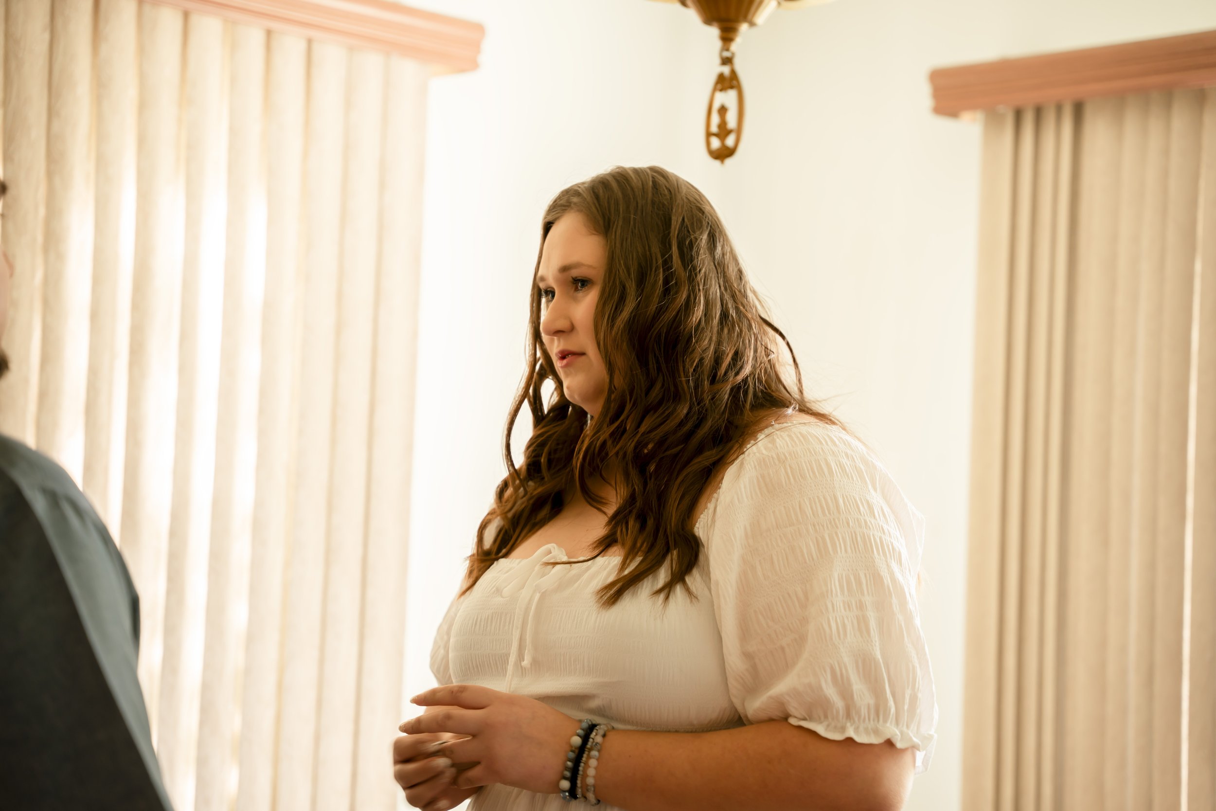 A woman with long, wavy brown hair and wearing a cream-colored blouse with textured fabric, standing indoors near a window with beige curtains, looking to her left with a serious or concerned expression.