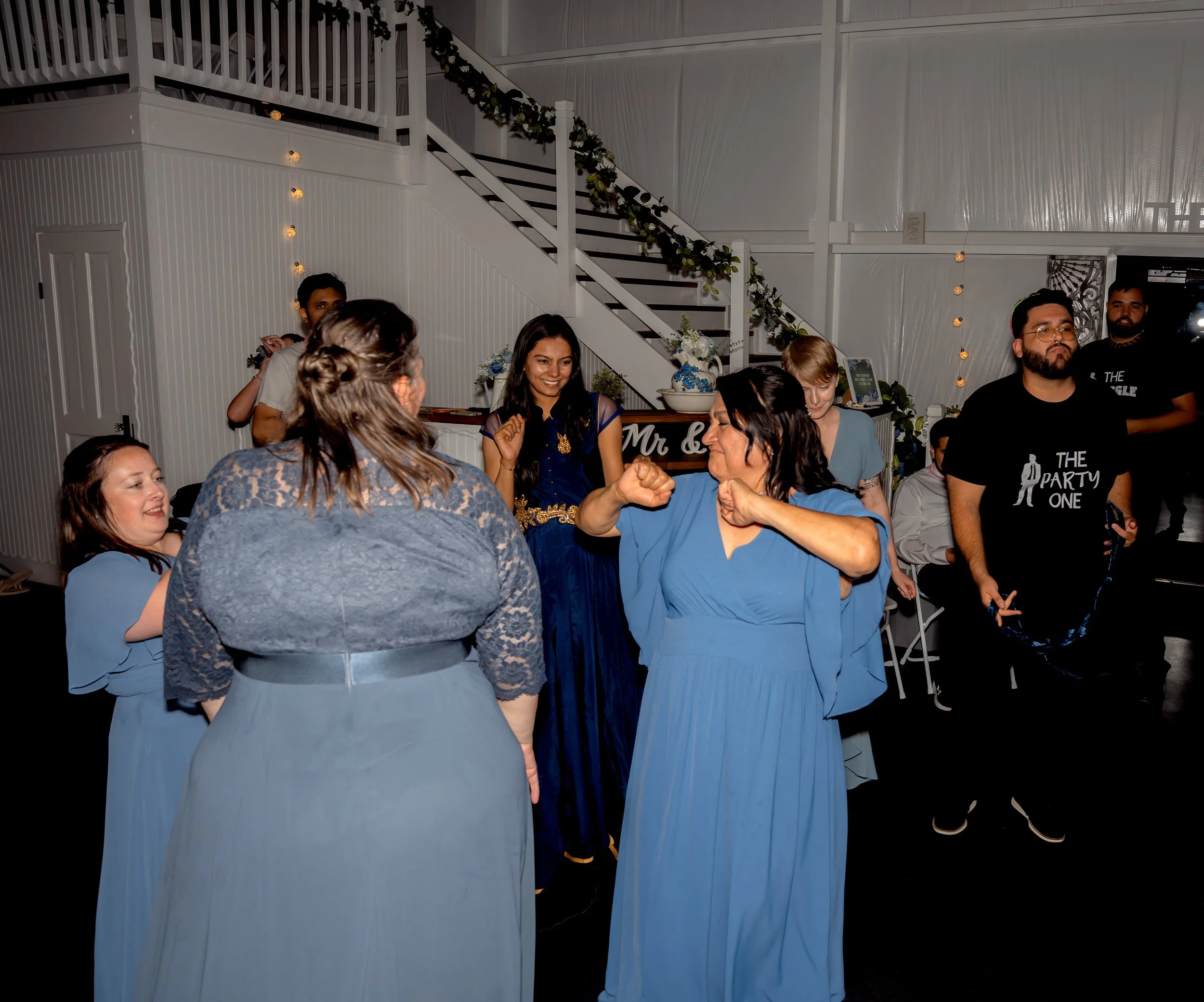 People dancing and enjoying at a celebration in a decorated indoor venue with a staircase and string lights.
