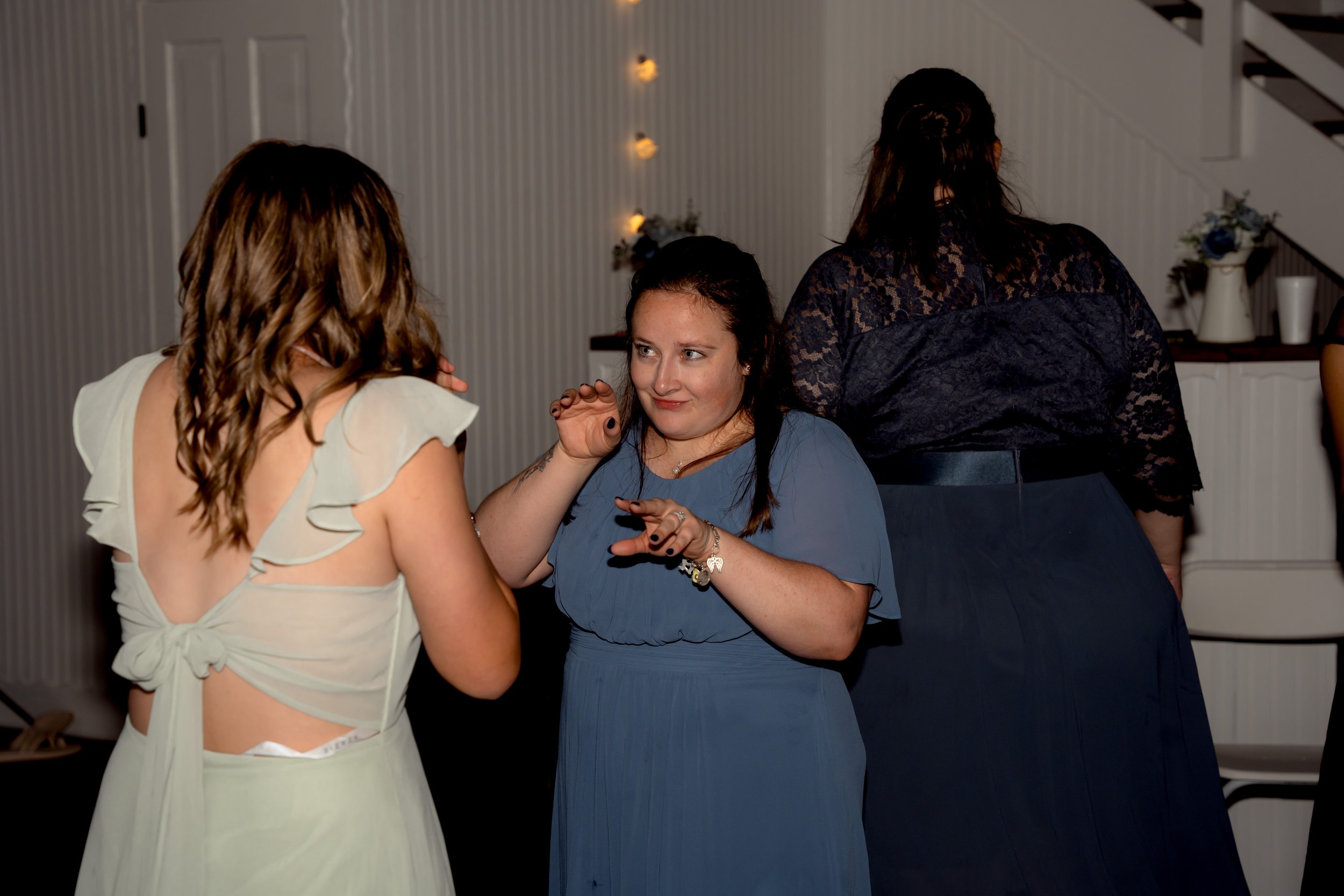 Three women engaged in conversation at an indoor event, with one woman in a white dress with open back, another in a blue dress facing her, and the third woman with her back to the camera in a black lace top.