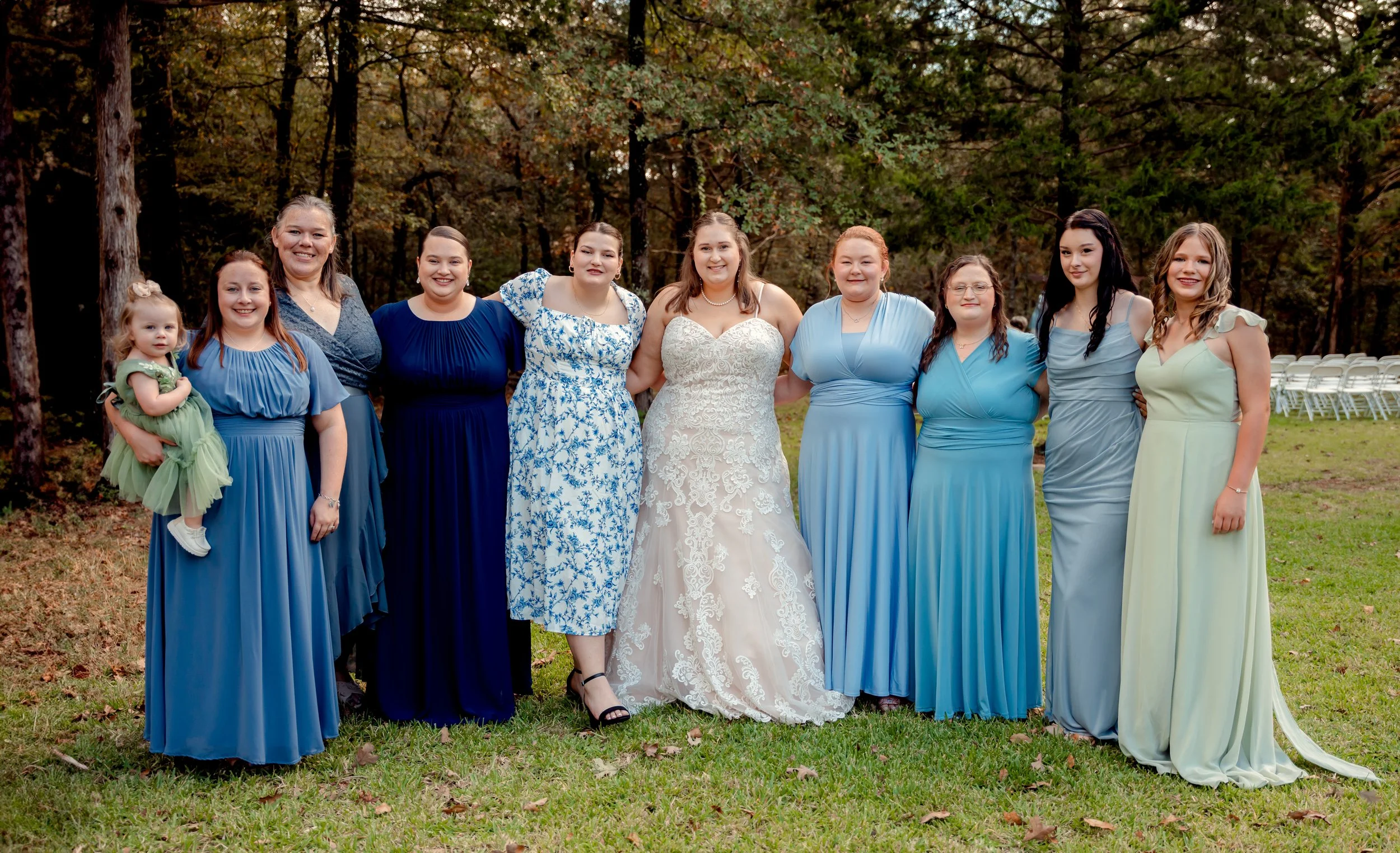 A group of women and a young girl dressed in formal attire standing outdoors on grass in a wooded area during daytime. The woman in the center wears a white wedding dress, surrounded by women in various shades of blue and pastel dresses, with empty c