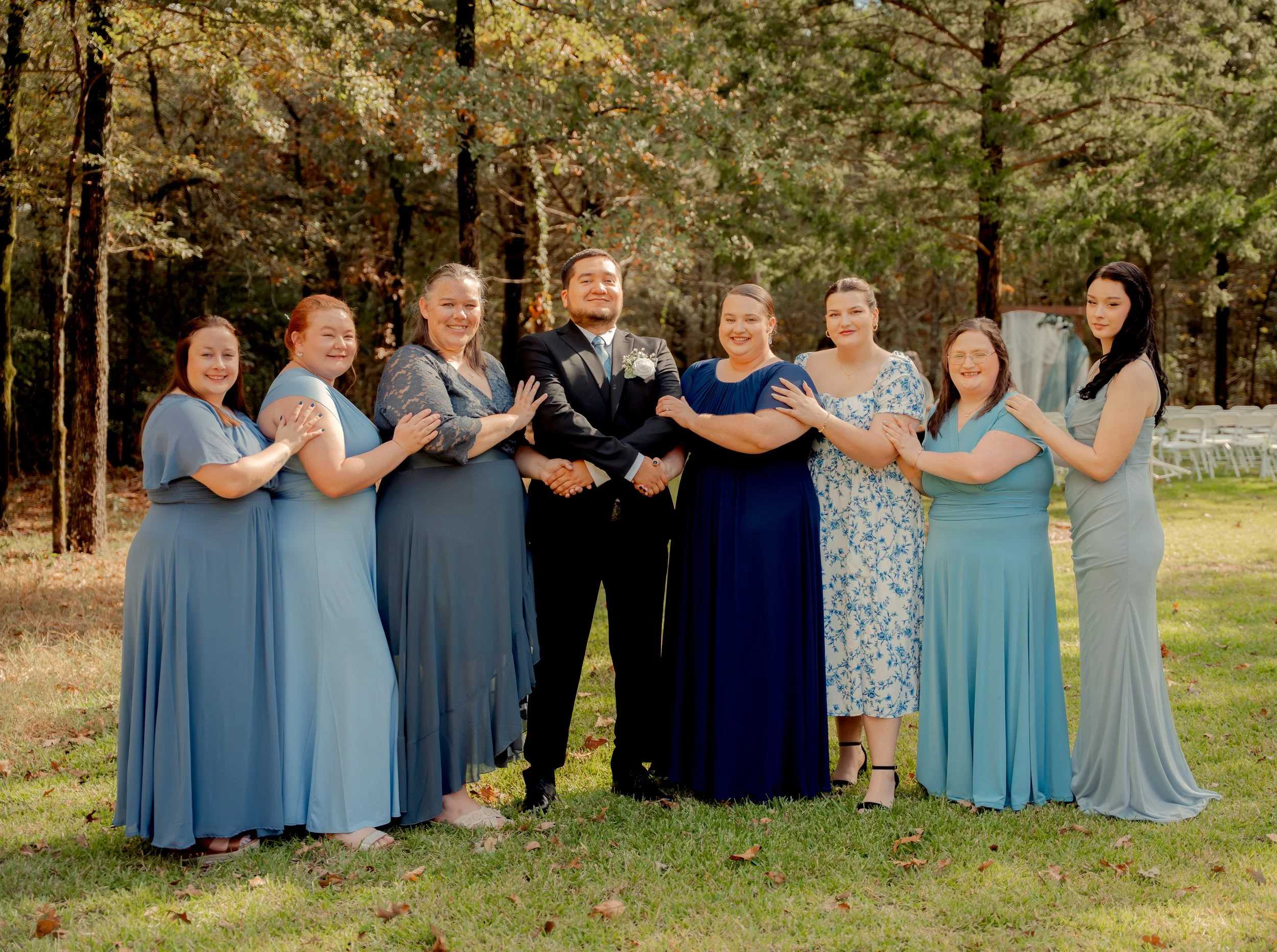 A group of nine people, including a groom and seven women in various shades of blue dresses, standing outdoors in a park with tall trees, posing for a photo at a wedding or special event.