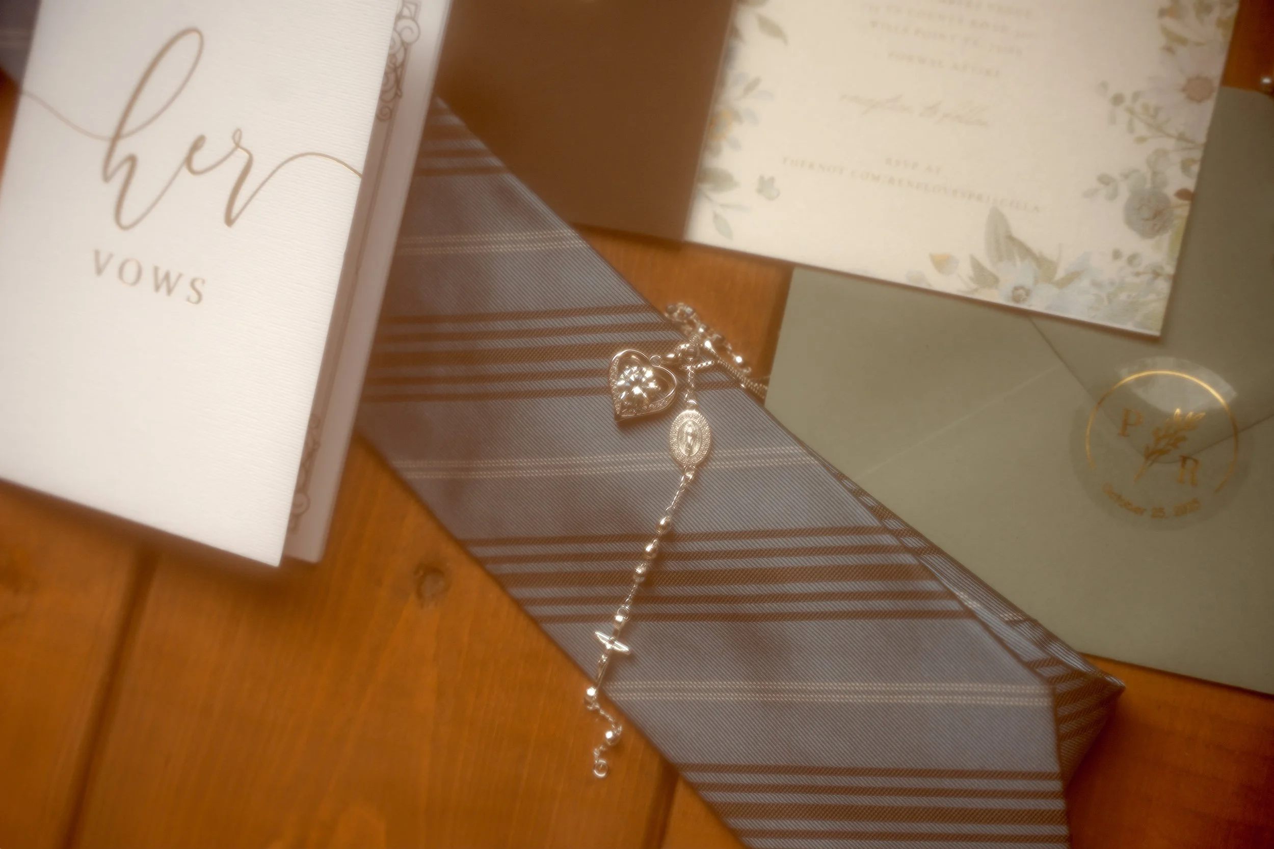 Silver bracelet with heart-shaped charm, prayer card with 'her vows' text, and wedding invitation with floral design on a table.