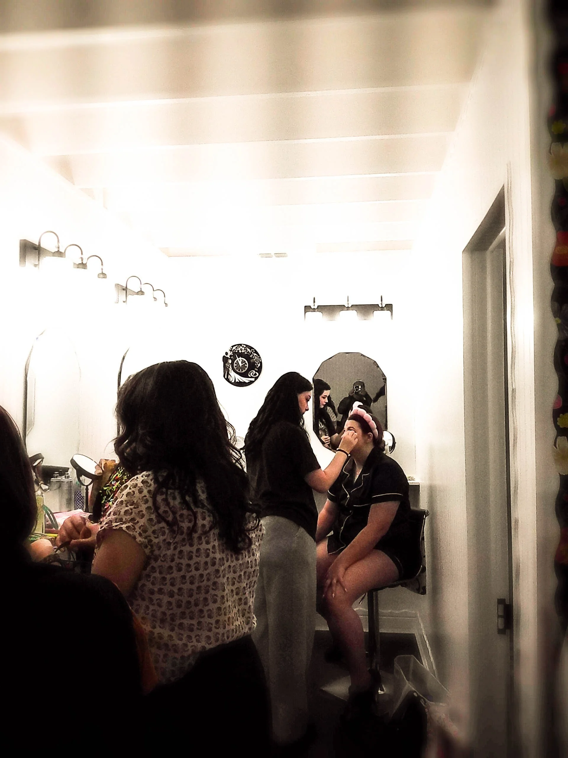 A makeup artist applying makeup to a woman sitting on a chair in a vanity room, with other women nearby and mirrors on the wall.