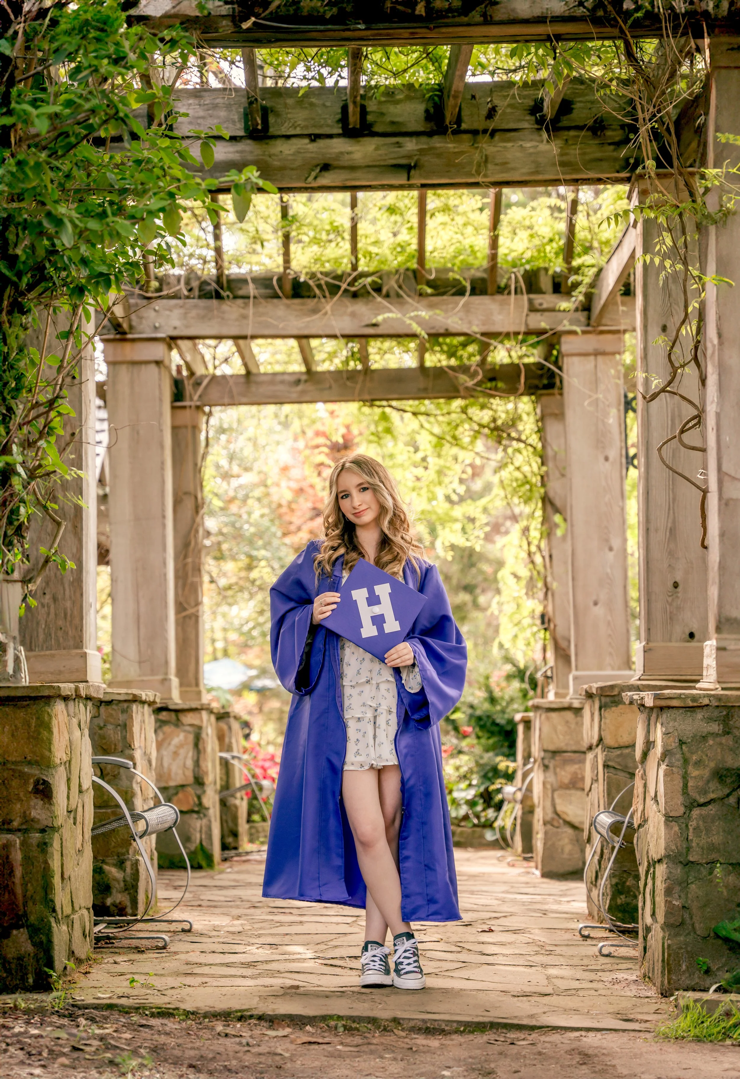 A young woman in a graduation gown and cap holding a diploma with a large 'H' stands on a stone and wooden walkway in a garden-like setting with lush greenery.