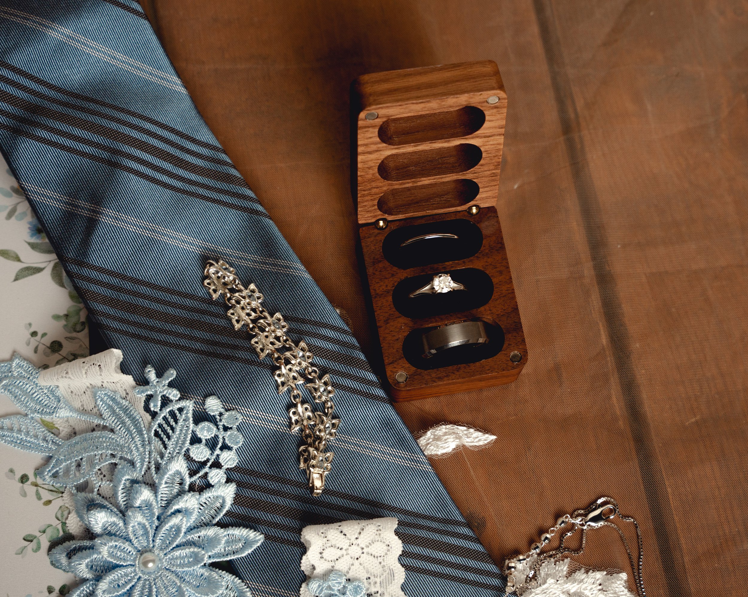 A wooden jewelry box containing rings, a brooch, and other jewelry, placed on a wooden surface next to a blue striped tie and lace floral accessories.