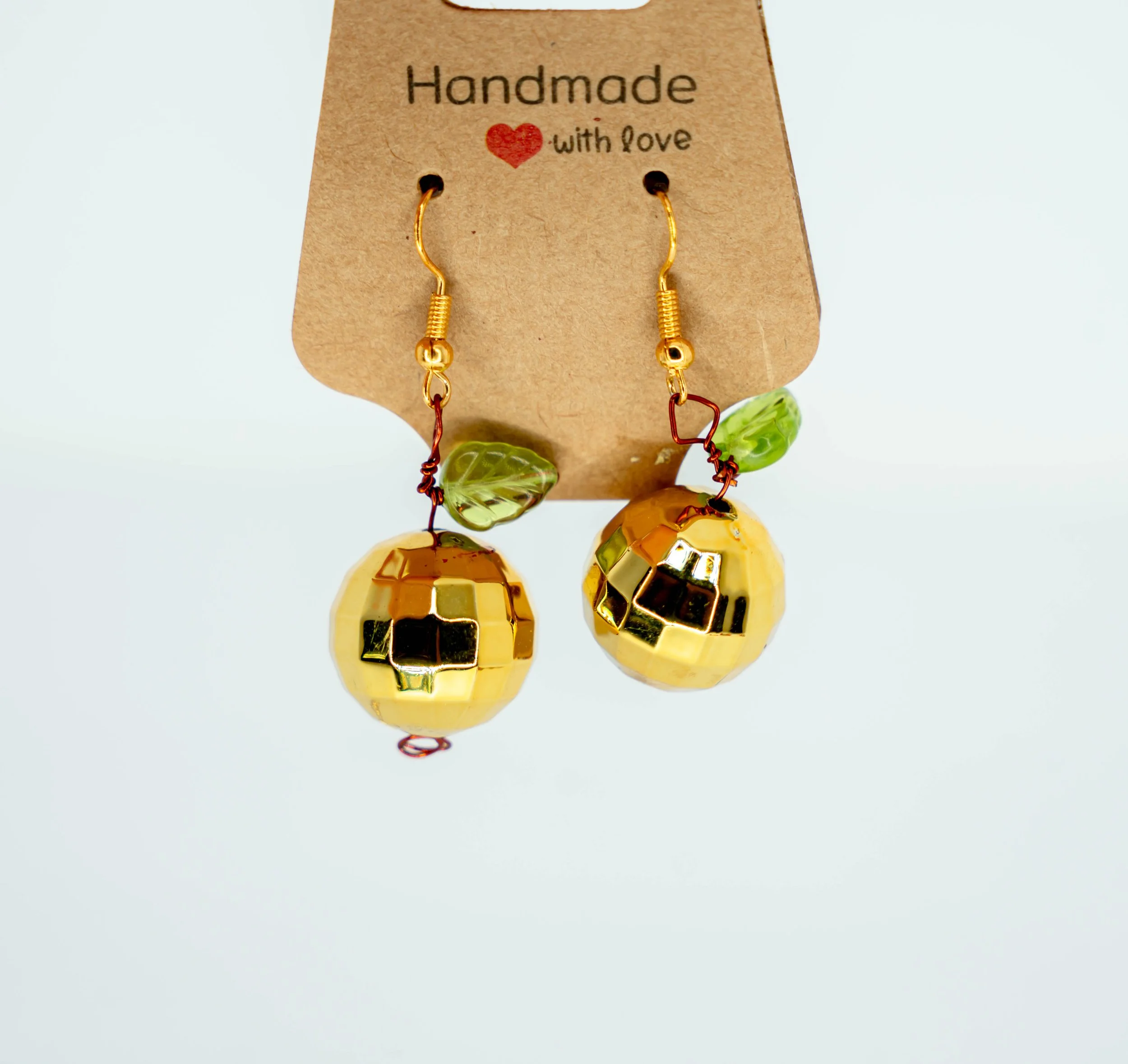 Handmade earrings with gold disco ball charms, green leaf beads, and red wire accents on a cardboard display that says 'Handmade with love'.