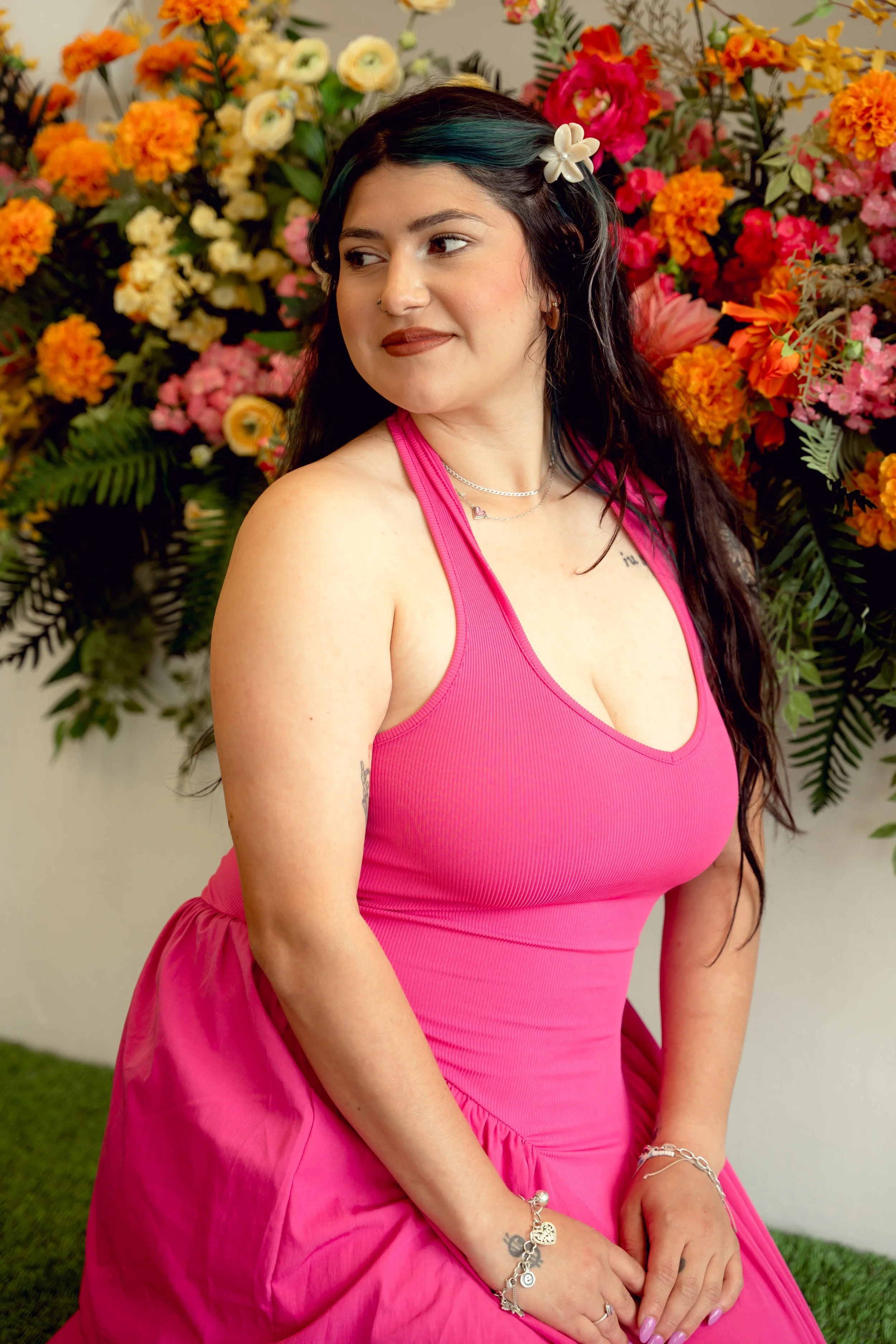 A woman with dark hair and a flower hair clip, wearing a pink dress, standing in front of a vibrant floral backdrop.