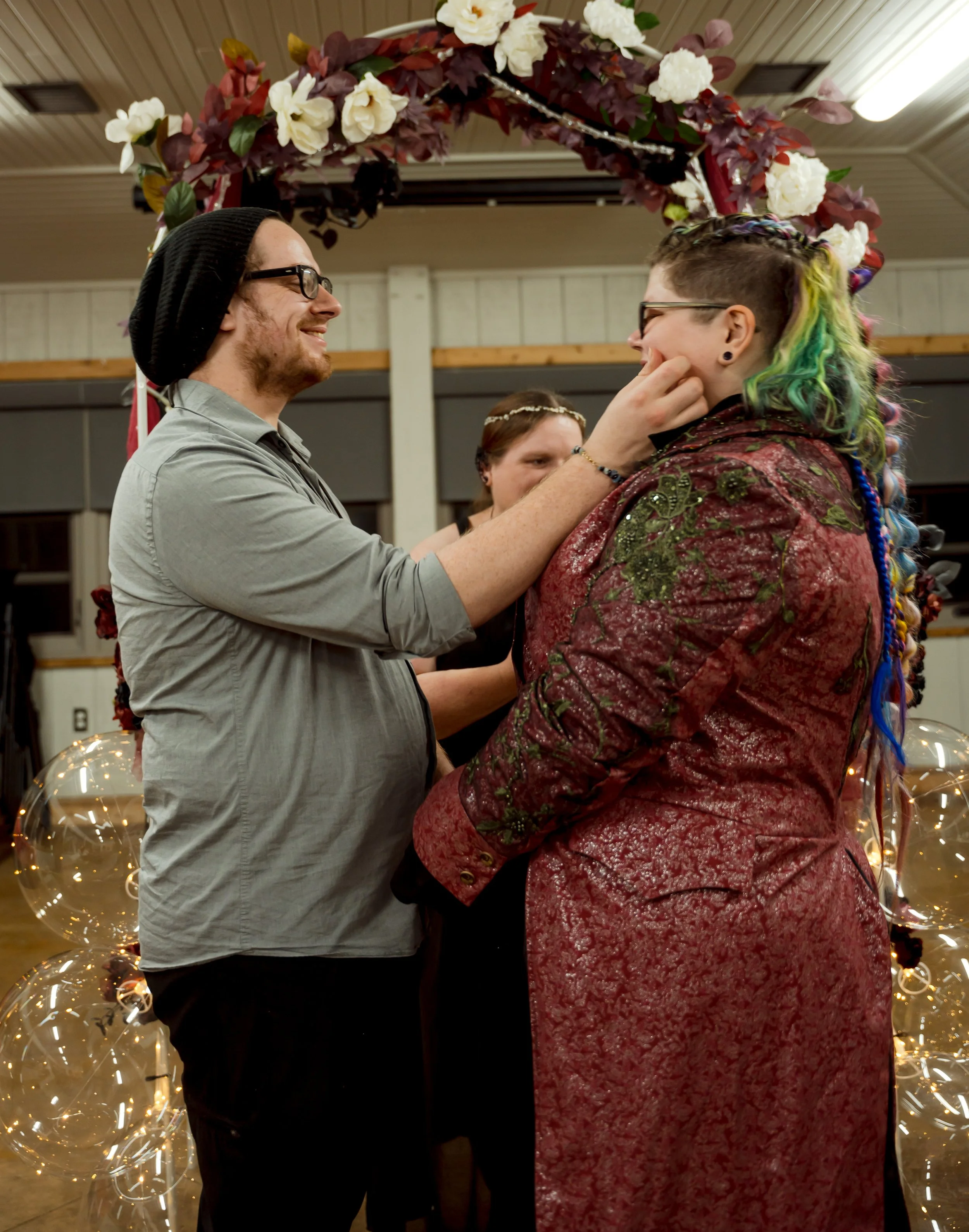 A person with rainbow-colored hair and glasses is being touched on the face by another person with glasses and a beanie, during a wedding or ceremony.