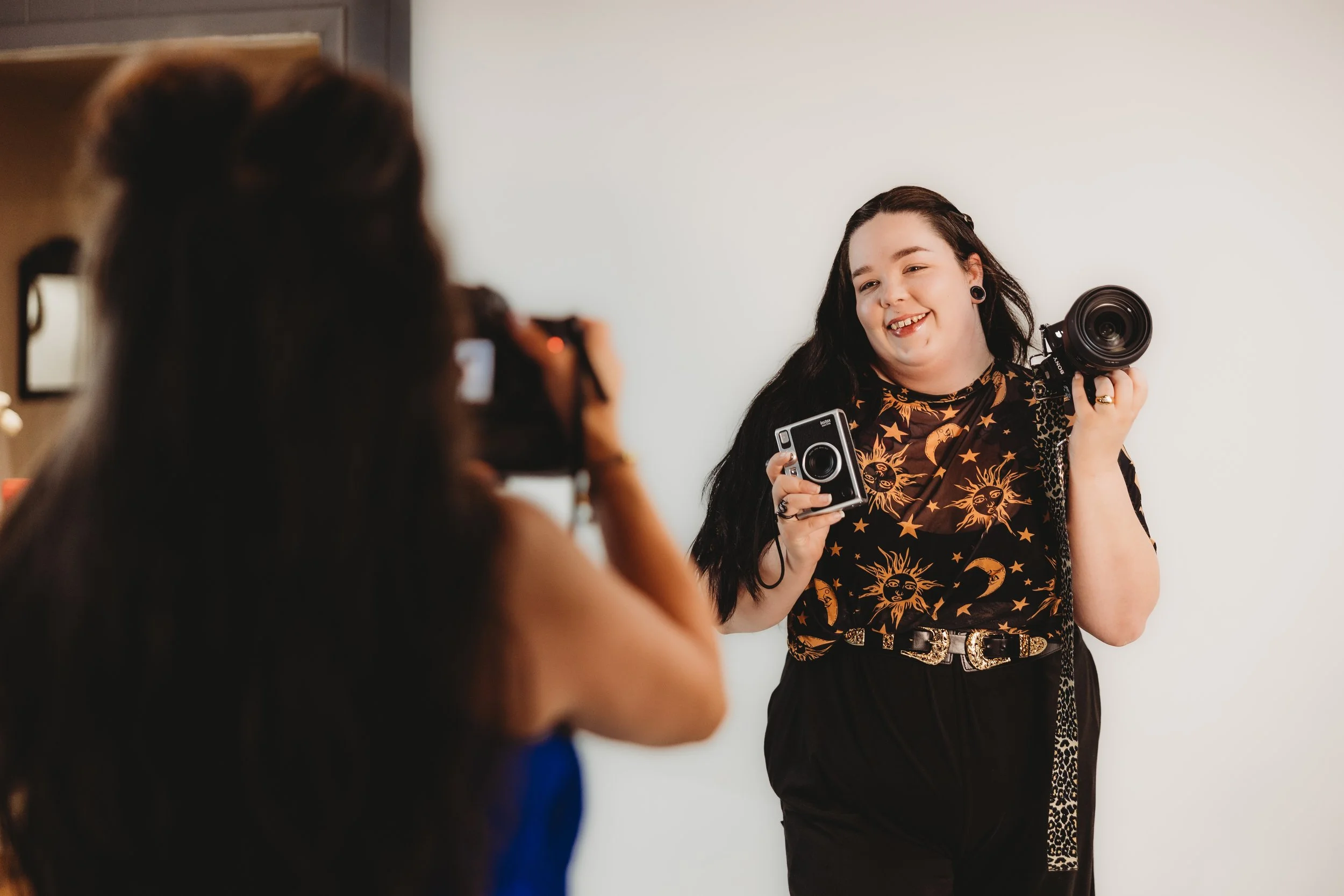 A woman with long dark hair, wearing a celestial-themed black and gold shirt, holding a camera and smiling, is posed for a photograph against a plain white background. Another woman, blurred in the foreground, is taking her picture with a camera.