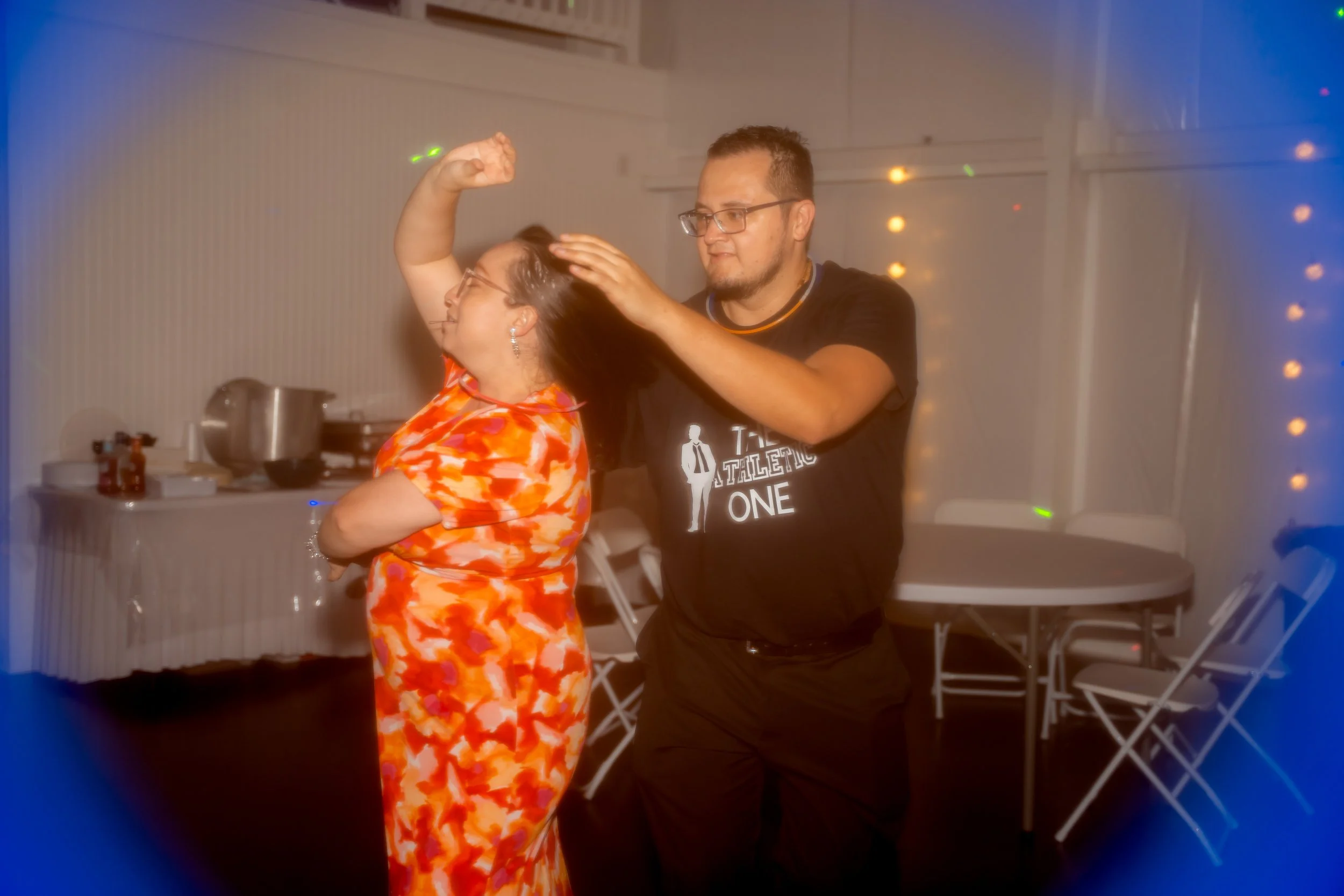 A man in glasses and a black T-shirt dances with a woman in an orange patterned dress at a party or celebration, with string lights in the background.