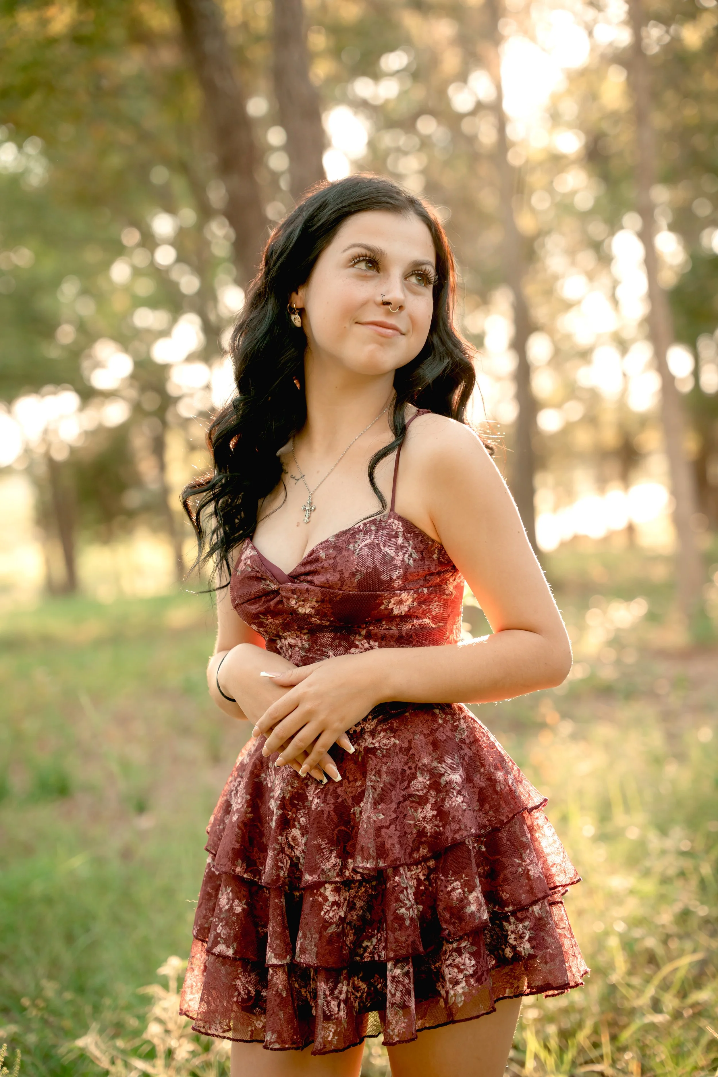 A young woman with long dark hair and light skin, wearing a floral burgundy dress, standing outdoors in a sunlit forest.