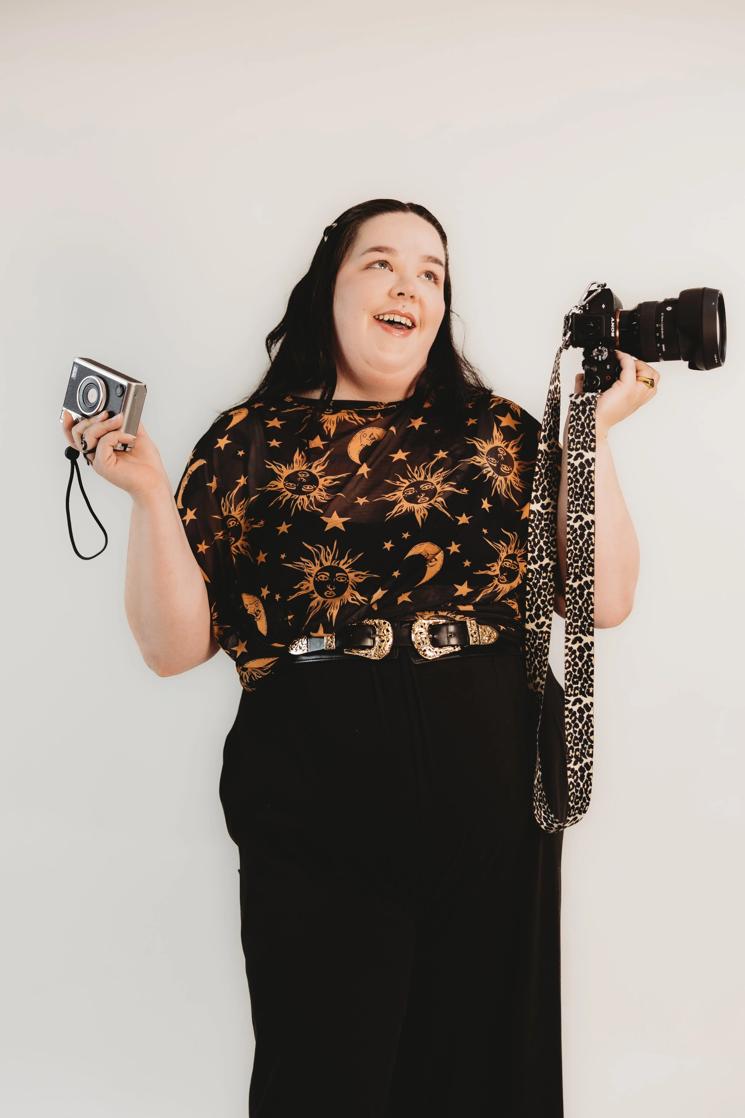 A woman standing against a white background, holding a point-and-shoot camera in her right hand and a larger DSLR camera with a leopard-print strap in her left hand. She has long dark hair and is wearing a black top with celestial patterns, black pants, and a decorative belt.
