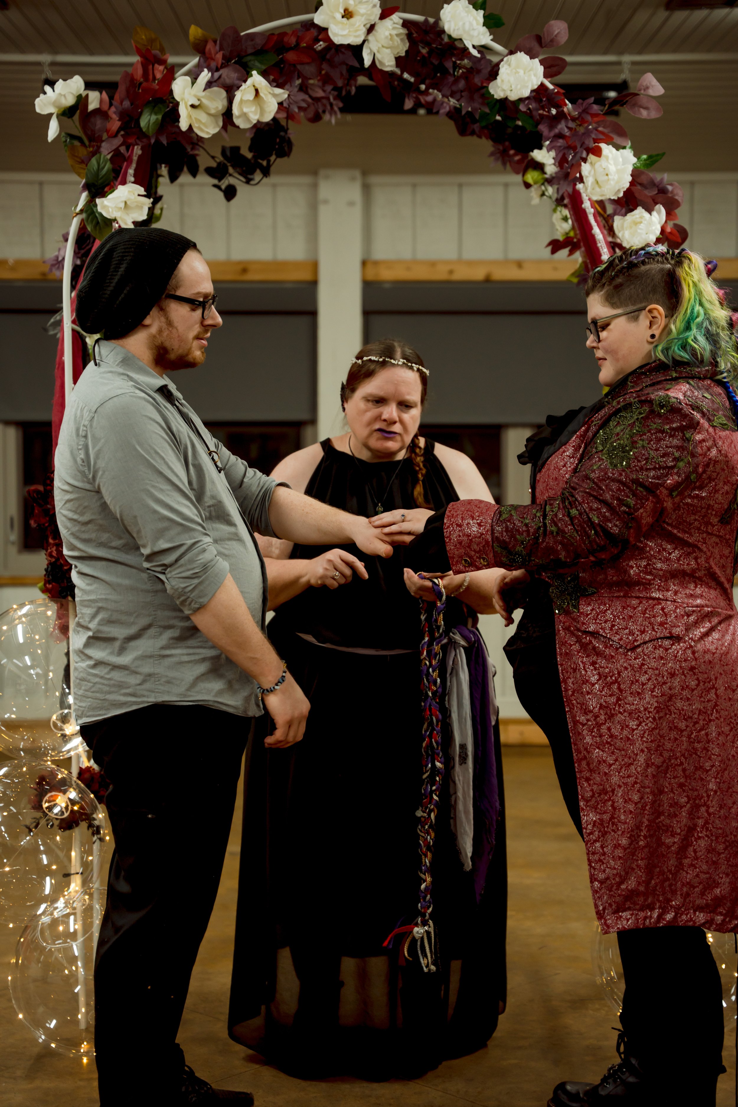 A couple getting married during a ceremony under a floral arch, officiated by a woman with braided hair, in a decorated indoor setting.