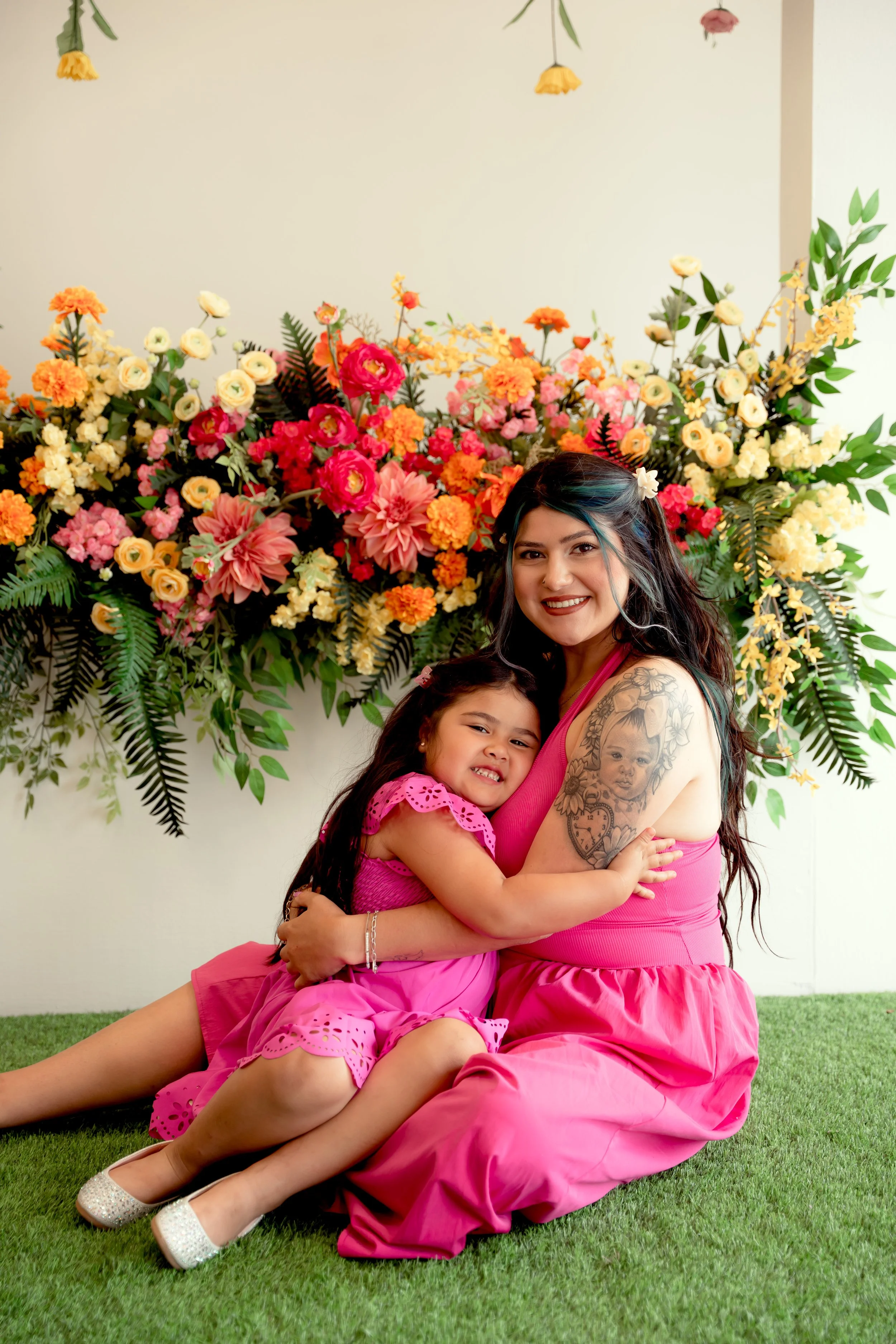 A woman and a young girl sitting on grass, hugging in front of a large colorful flower arrangement with pink, yellow, and orange flowers and green leaves, both smiling at the camera.