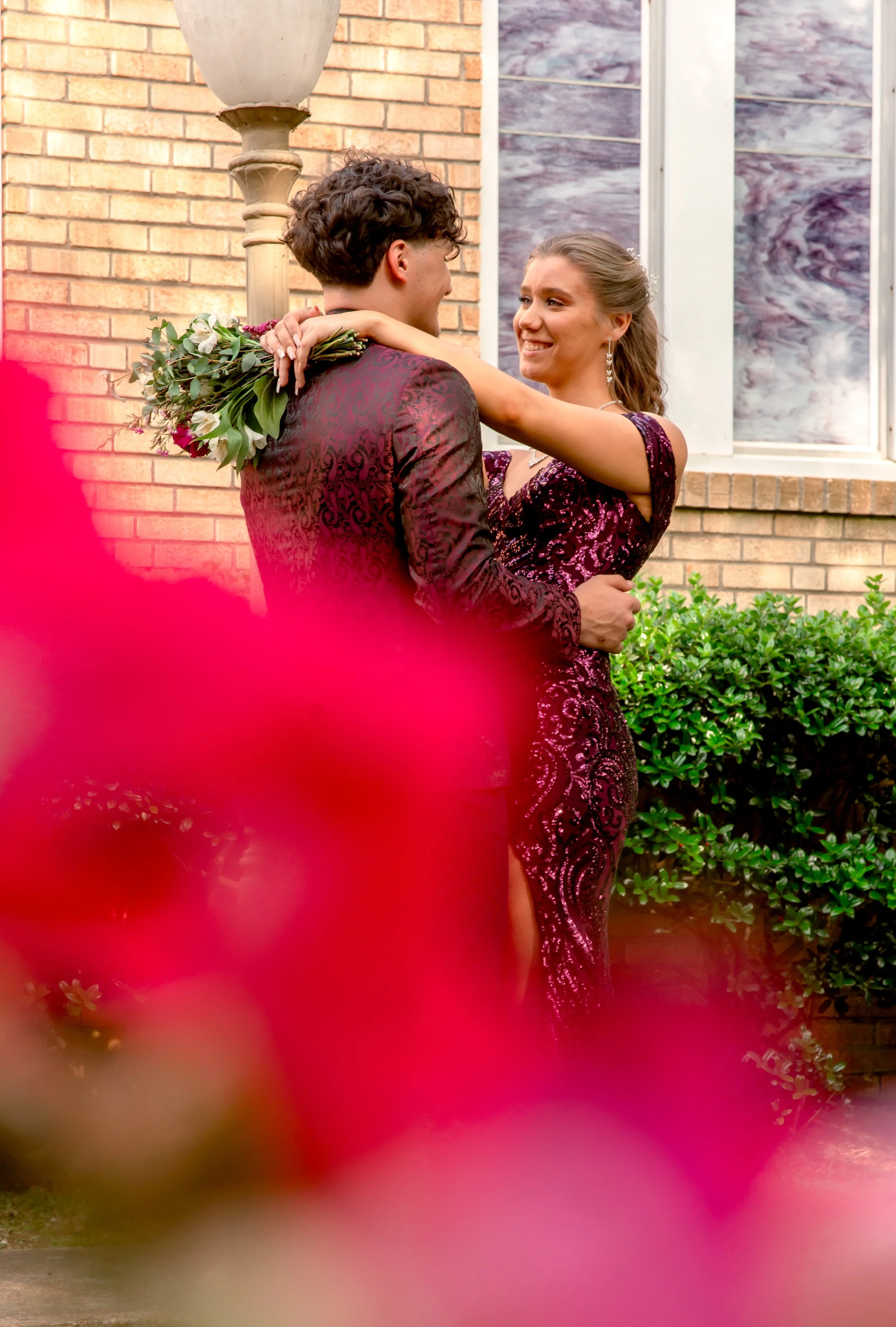 A couple in formal attire embraces outdoors in front of a brick building, with pink flowers out of focus in the foreground.