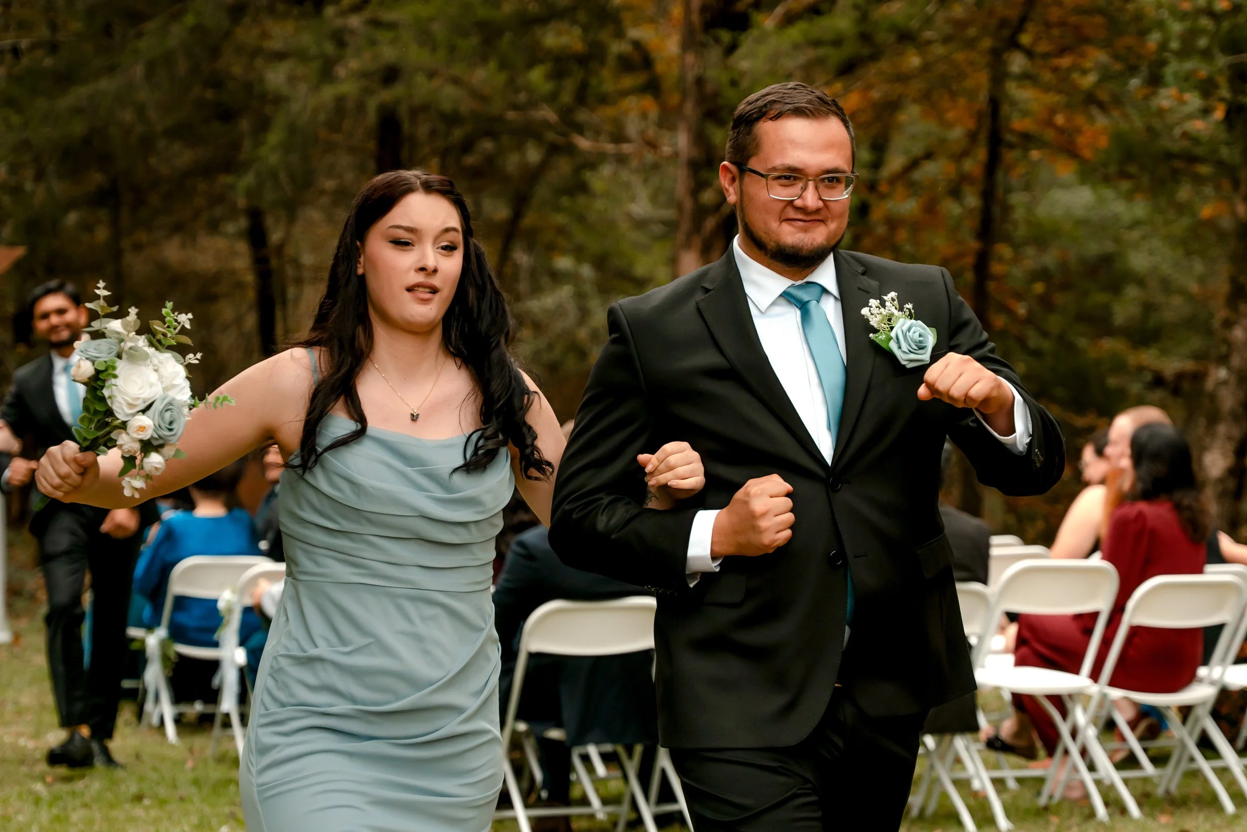 Couple running at outdoor wedding ceremony, woman holding bouquet, man in suit with boutonniere, guests seated in background.