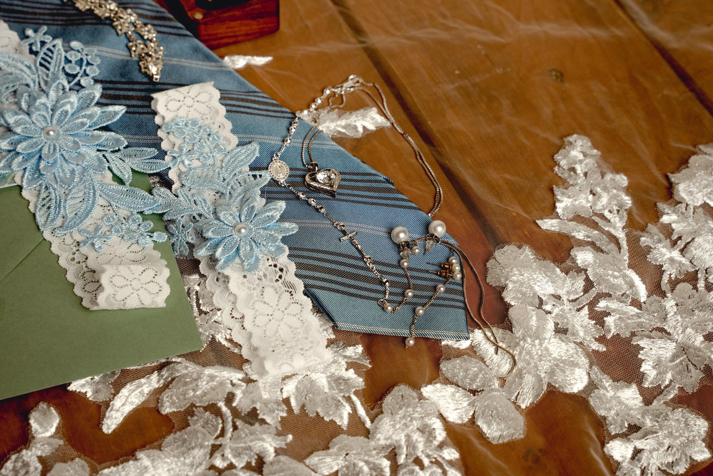 Shattered glass on a wooden surface with scattered jewelry including pearl necklaces, a heart-shaped necklace, and lace fabric with blue floral embroidery and lace trim.
